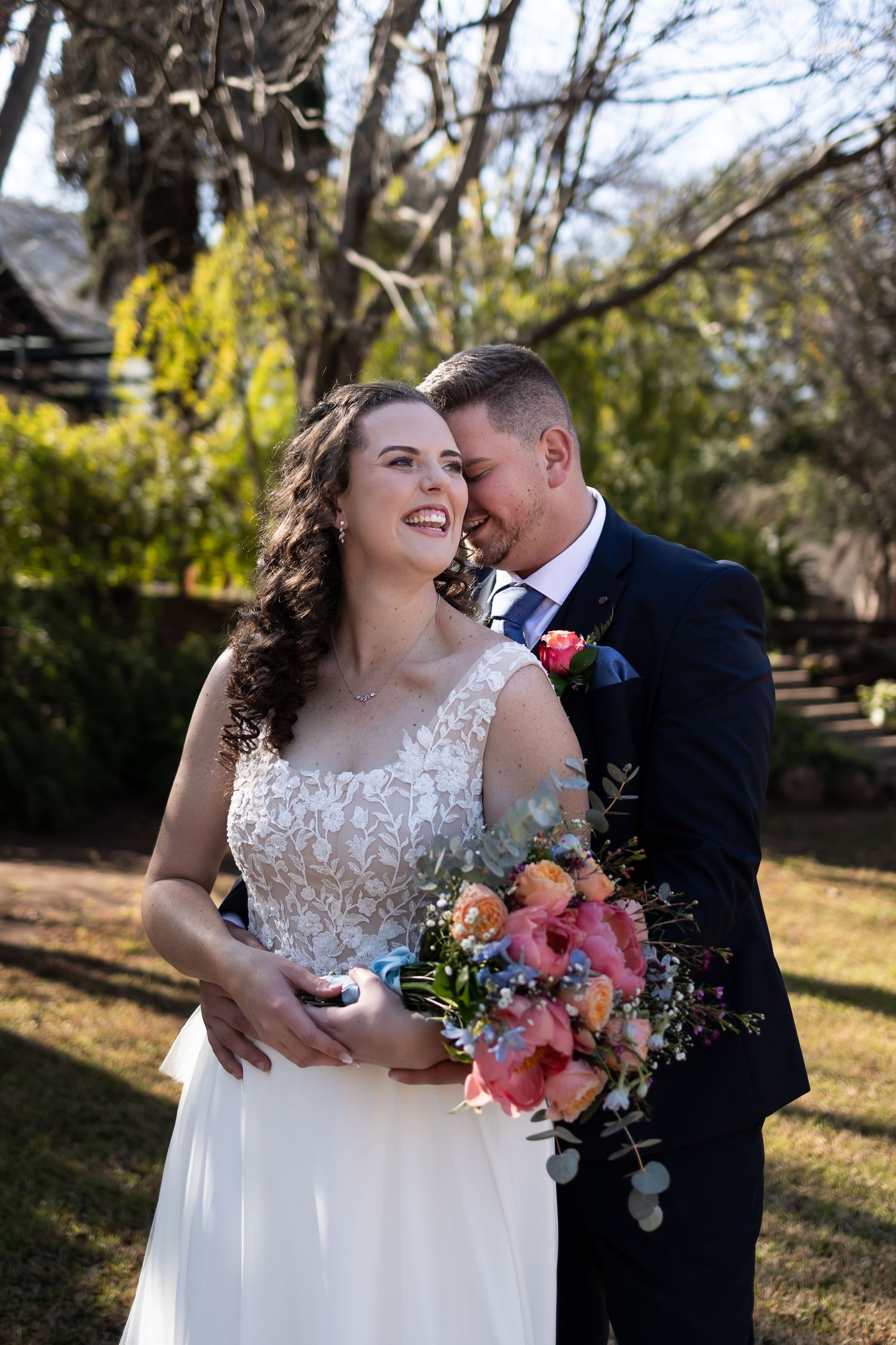A newlywed couple shares tender moments outdoors in a garden setting with the bride holding a colorful bouquet.