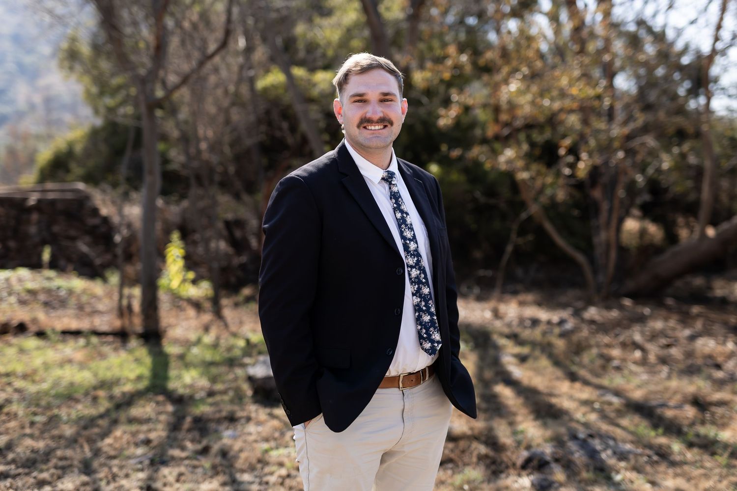Professional outdoor portrait of someone in a black blazer and patterned tie standing against an autumn background.