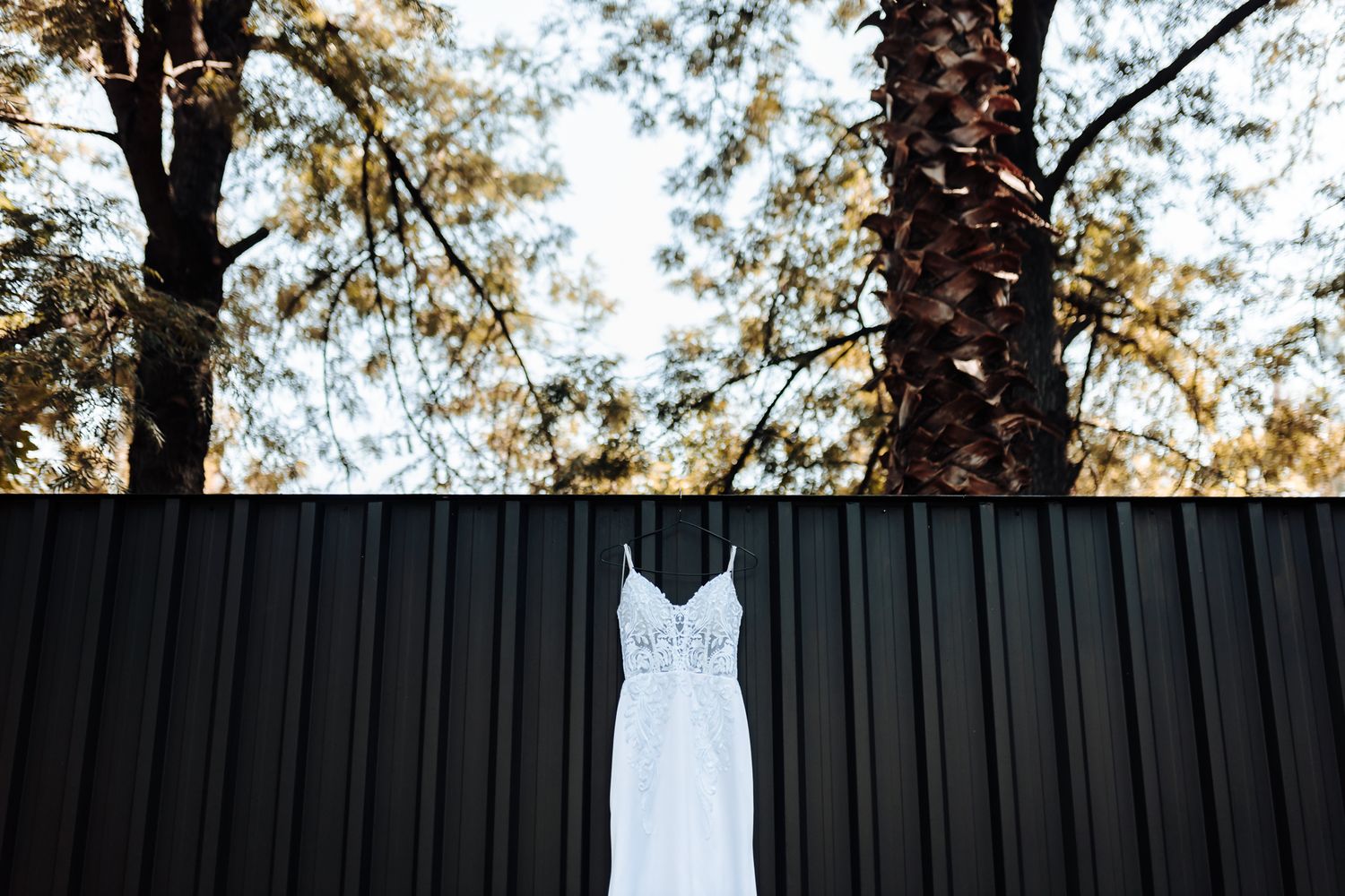 White wedding gown suspended against black metal wall with trees visible in background.