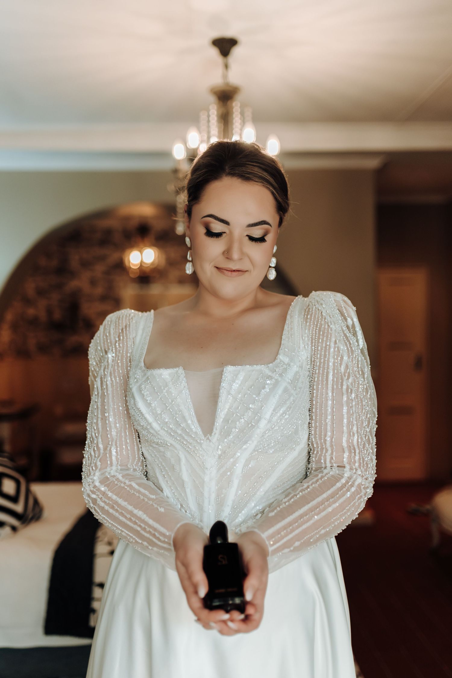 Wedding dress detail shot focusing on square neckline and sheer beaded sleeves under chandelier.