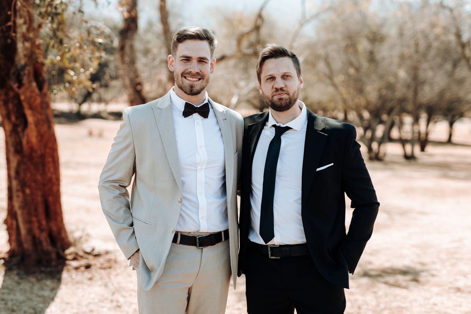 Two men in contrasting formal suits pose together at an outdoor wedding venue.