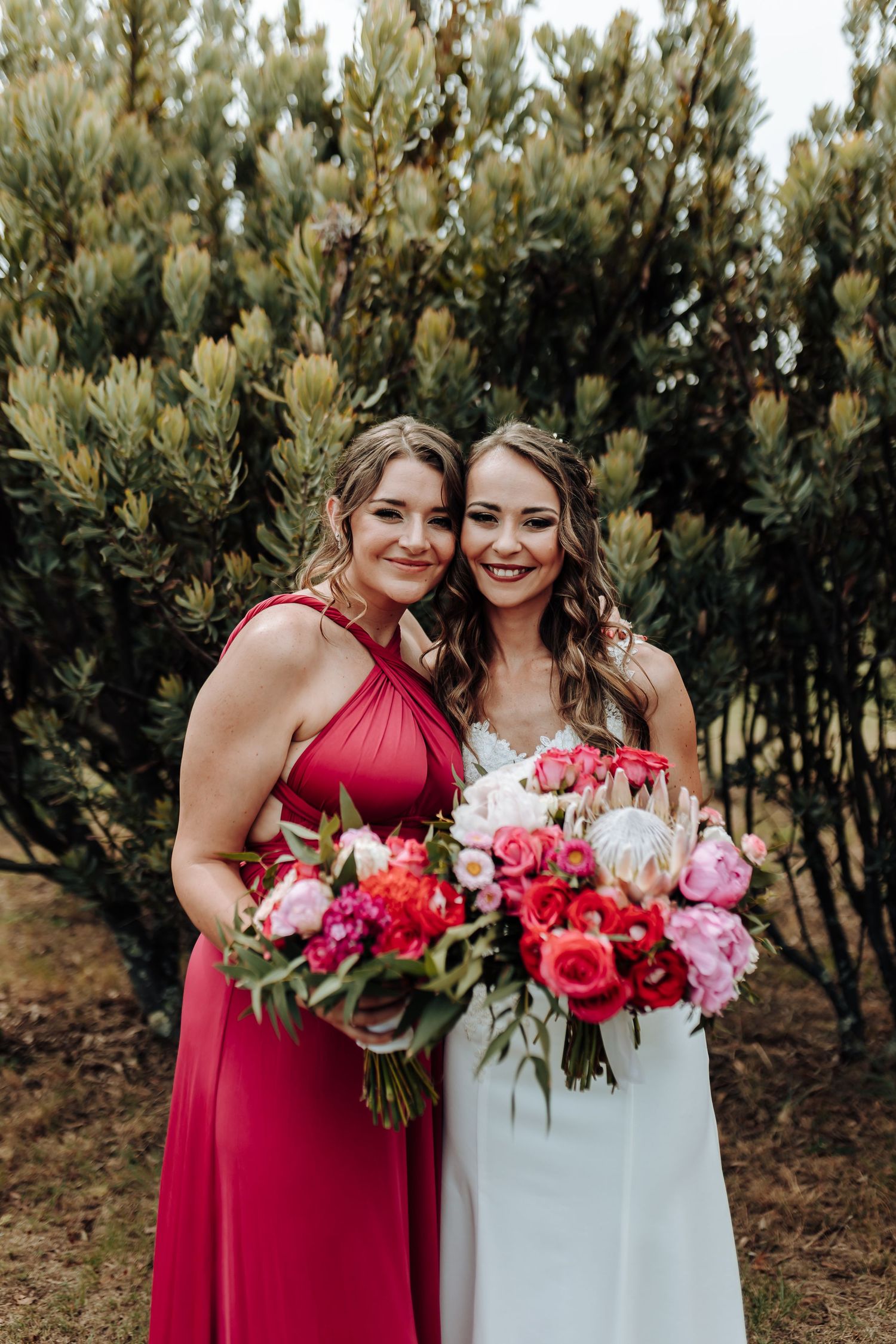 Bridesmaid in red dress and bride in white pose together with vibrant pink and red bouquets.
