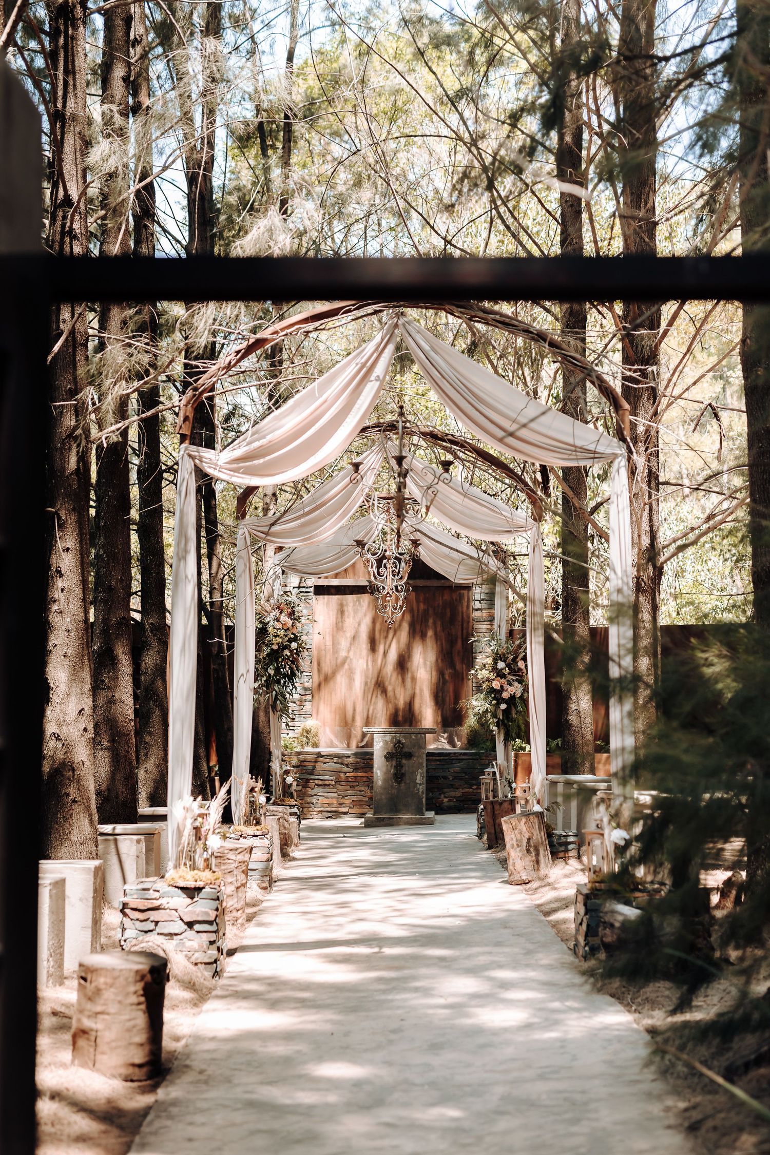 Romantic outdoor wedding ceremony aisle decorated with draped fabric and lanterns in a forest setting.