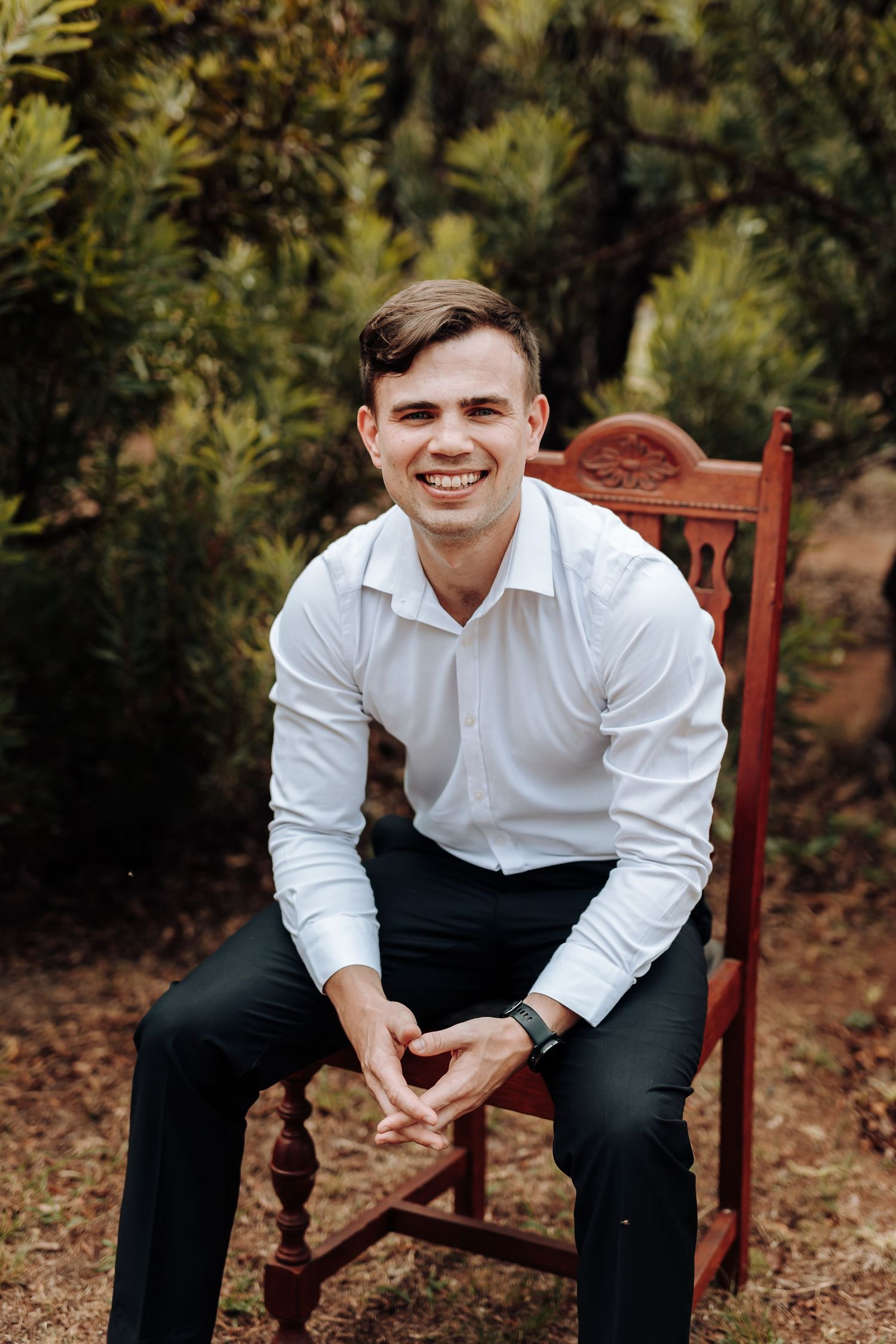 A seated figure in white dress shirt and dark pants outdoors among greenery.