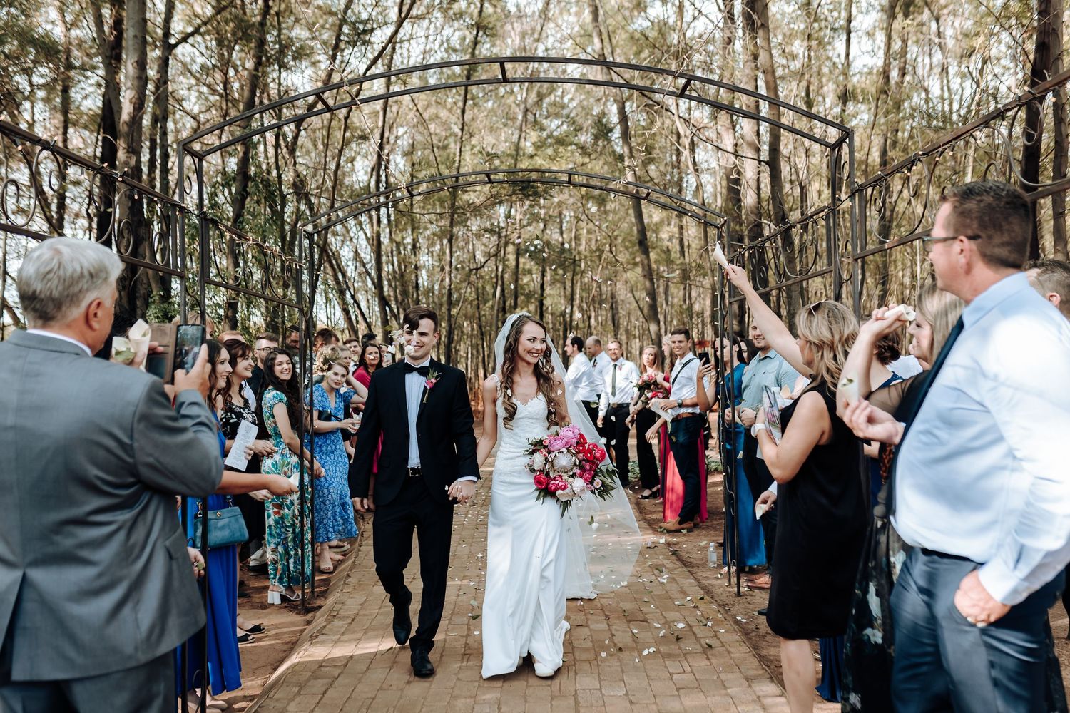 Wedding processional walking through wooded outdoor venue with guests watching from either side.