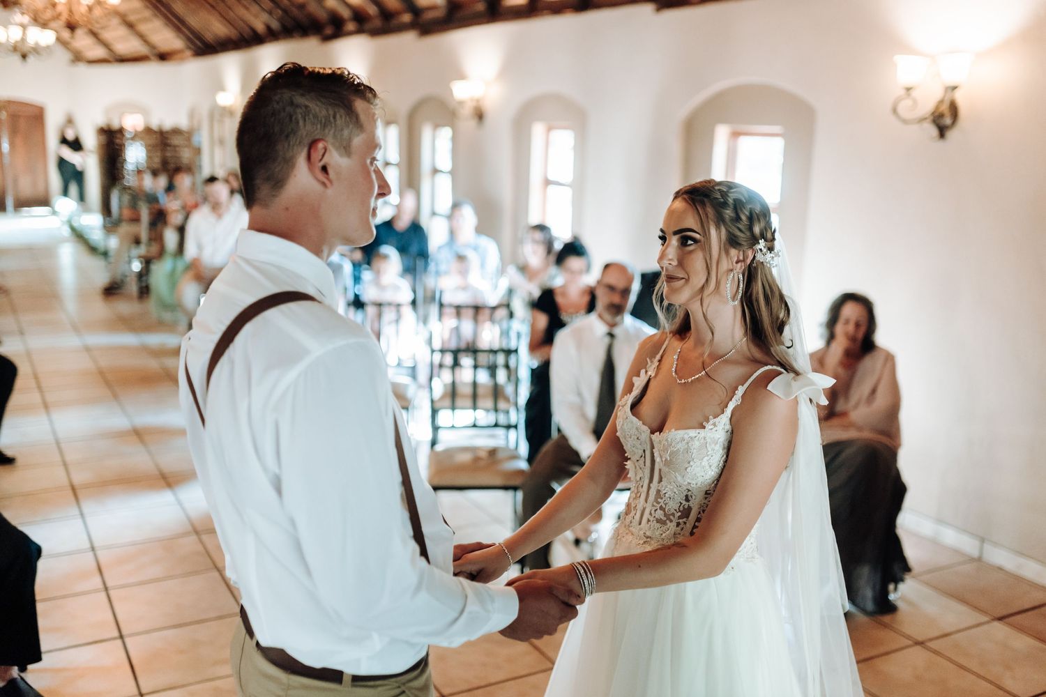 Couple holds hands during wedding ceremony in rustic venue with terra cotta floors.
