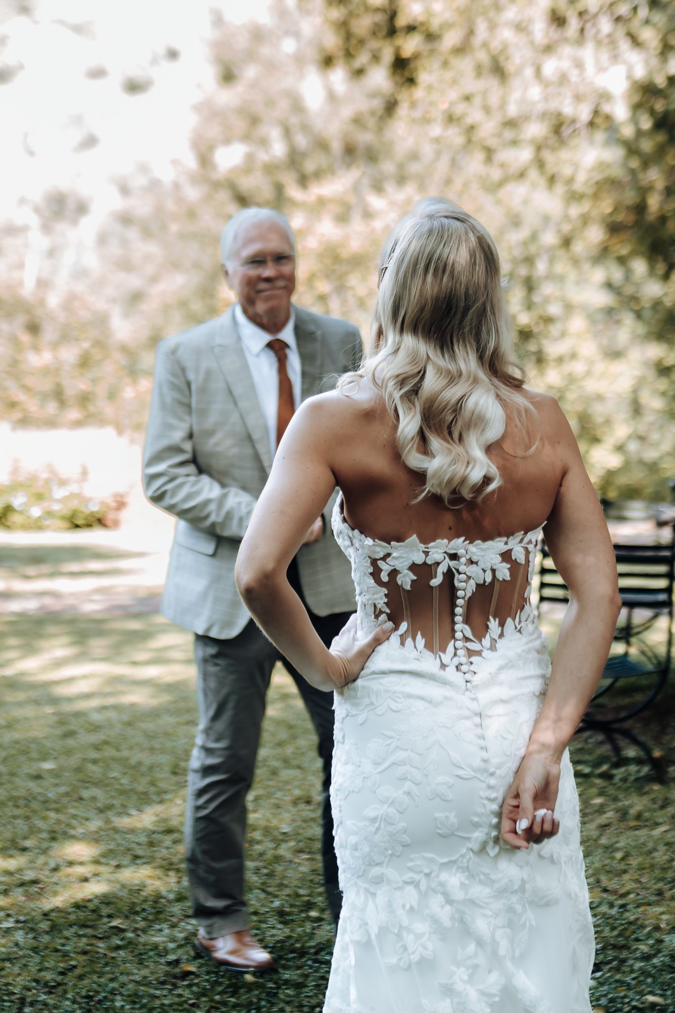 Back view of bride in sheer lace dress standing with man in light suit outdoors.