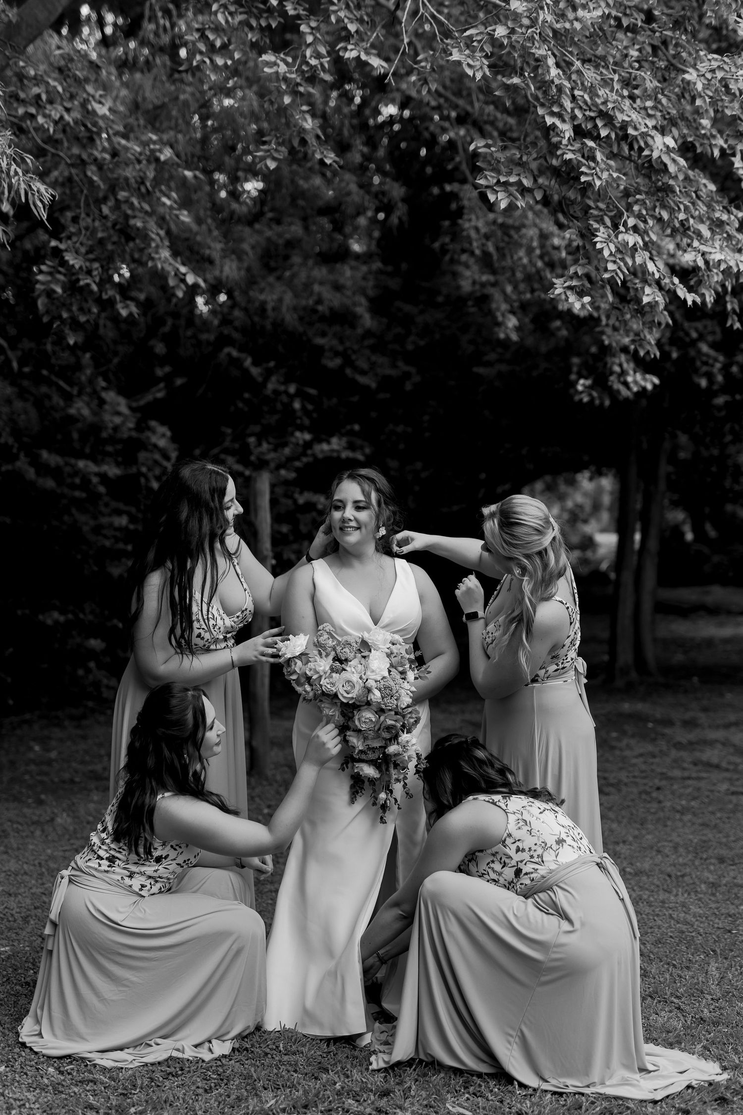 Black and white photo of bridal party in elegant dresses gathered for candid moment outdoors.