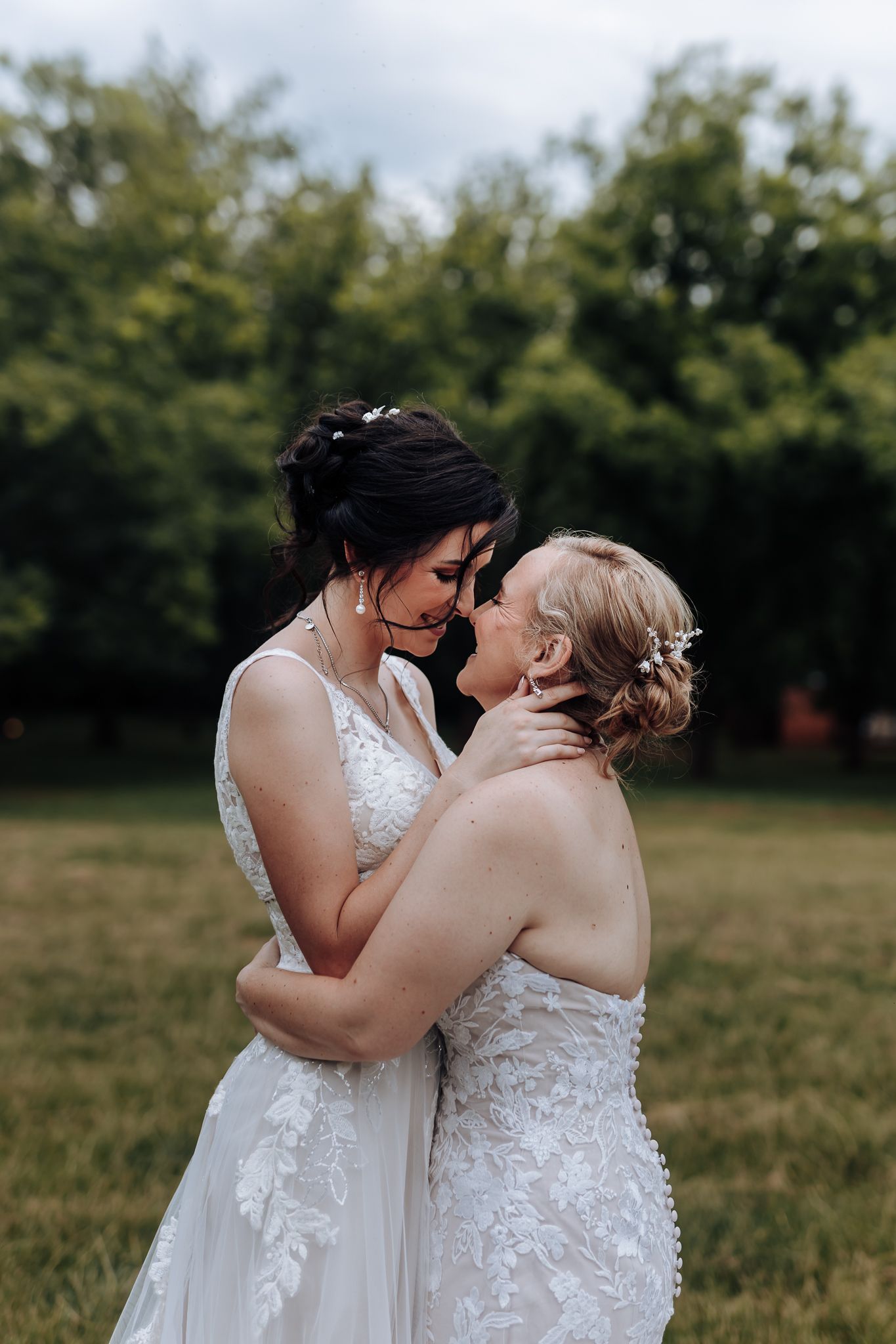 Two people in white wedding gowns walking hand in hand through a tree-lined park.