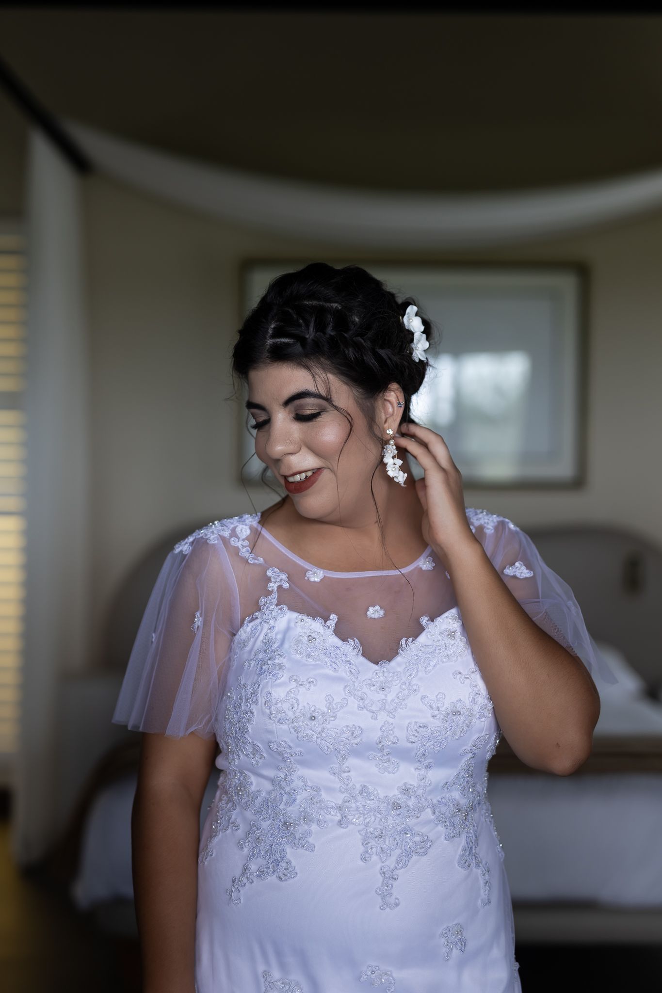 A bride wearing a sheer-sleeved white lace wedding dress adjusts her earring.