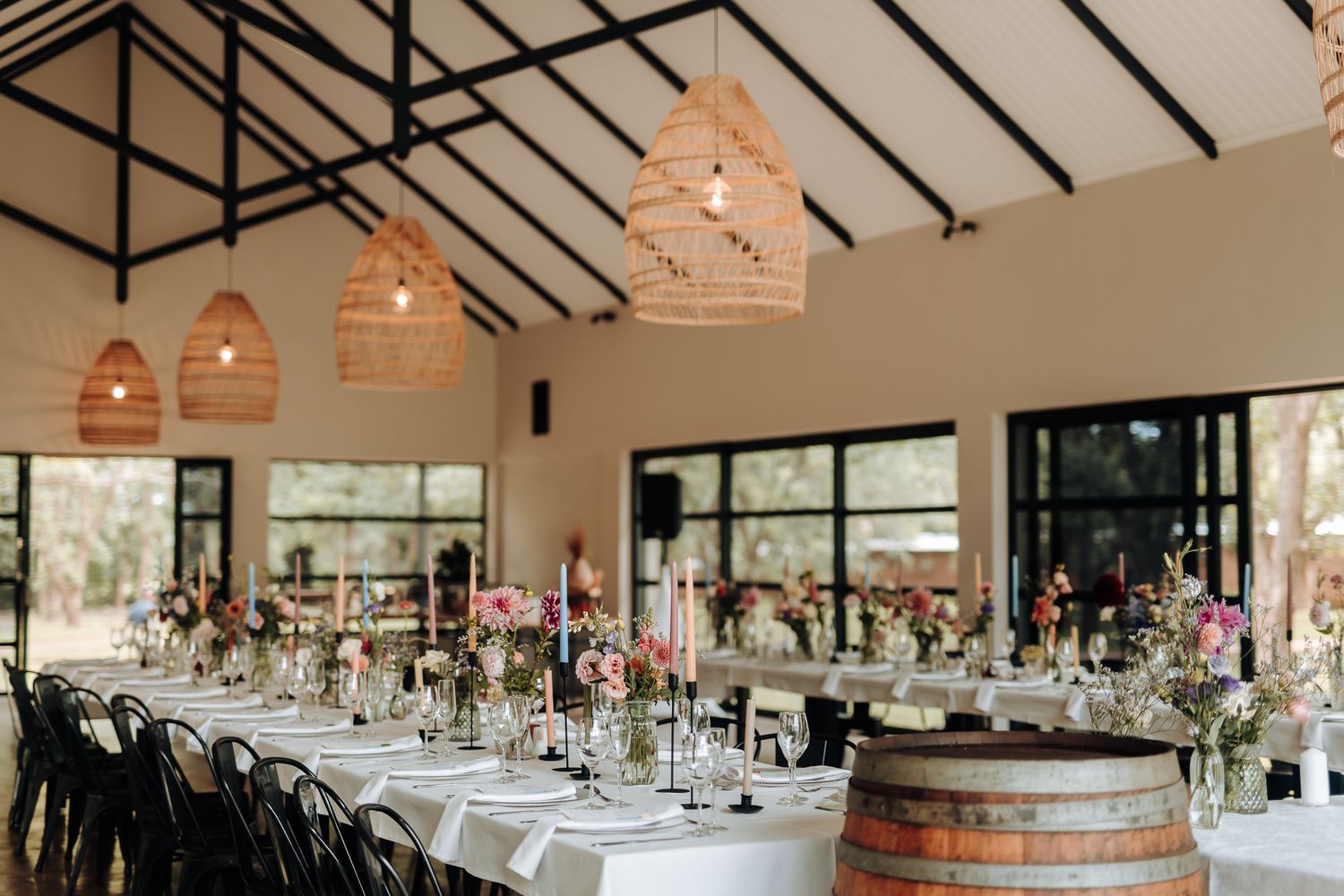 Modern reception hall with long dining tables, black chairs, and rustic wooden barrel accents.