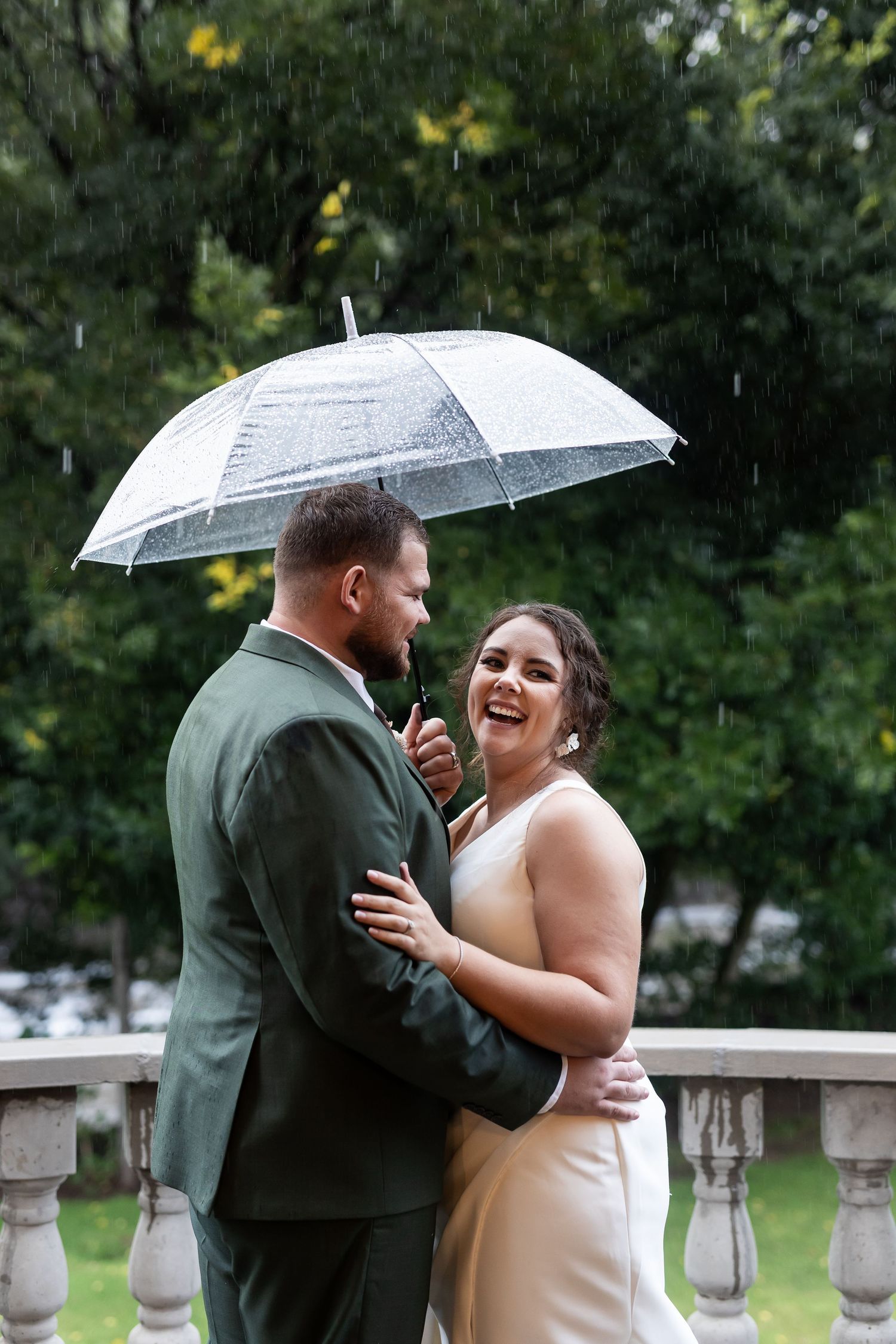 Couple laughs together while sharing clear umbrella on stone balcony.