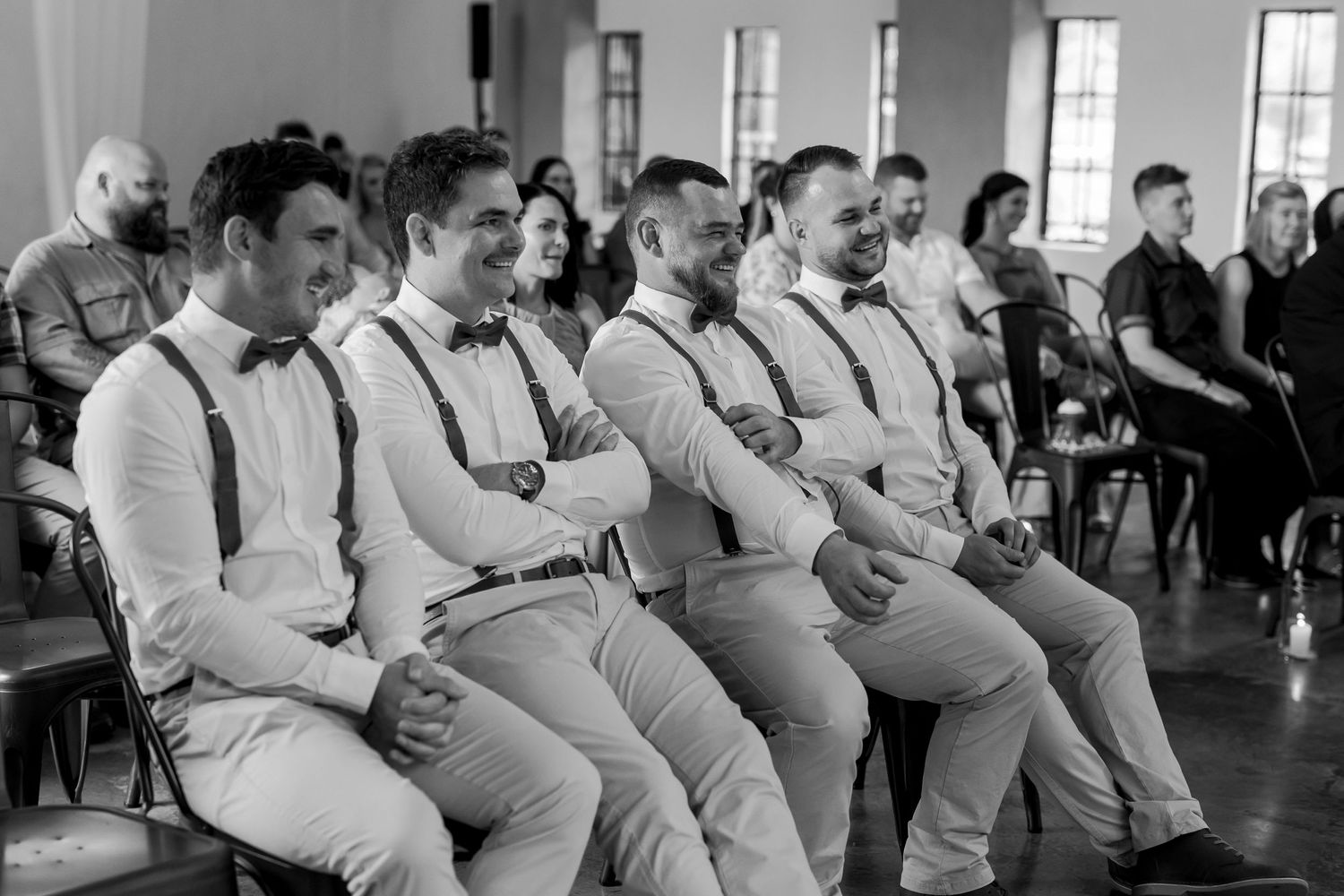 Groomsmen in light colored suits and suspenders sit together during a wedding ceremony.