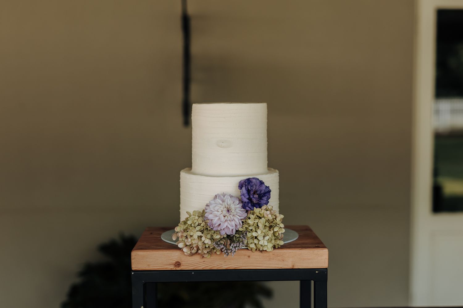Elegant white wedding cake decorated with purple flowers and greenery on wooden stand.
