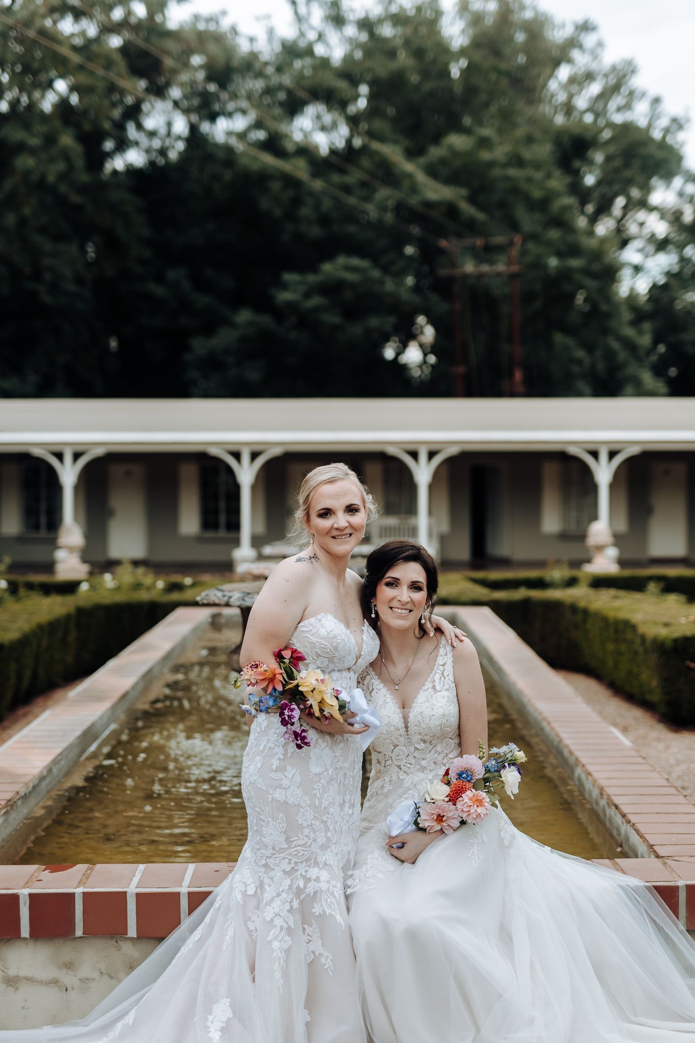 Two brides in lace gowns pose with bouquets by garden fountain at elegant venue.