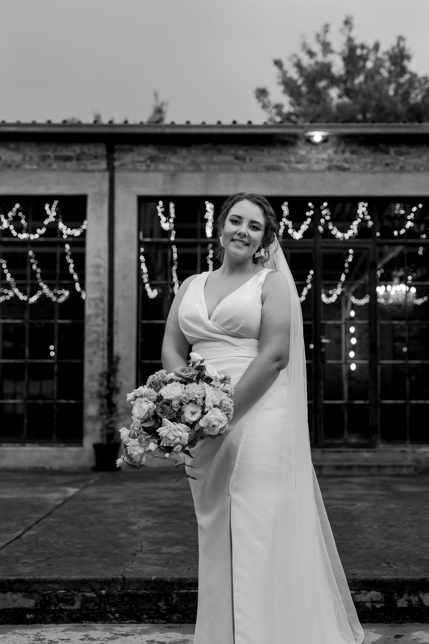 Black and white bridal portrait featuring elegant dress and bouquet against industrial backdrop.