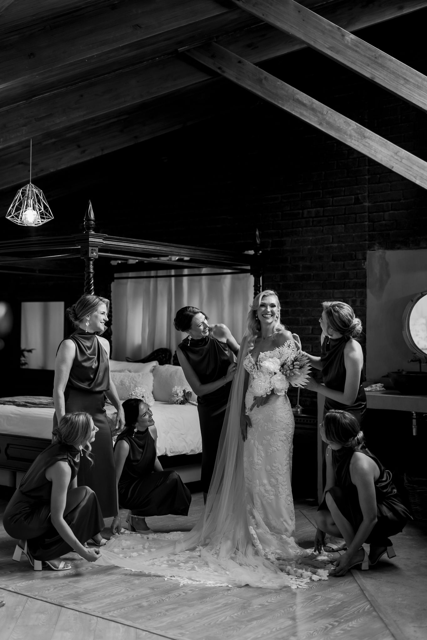 Wedding party helps with the bridal gown in a rustic indoor setting with exposed beams and brick walls.