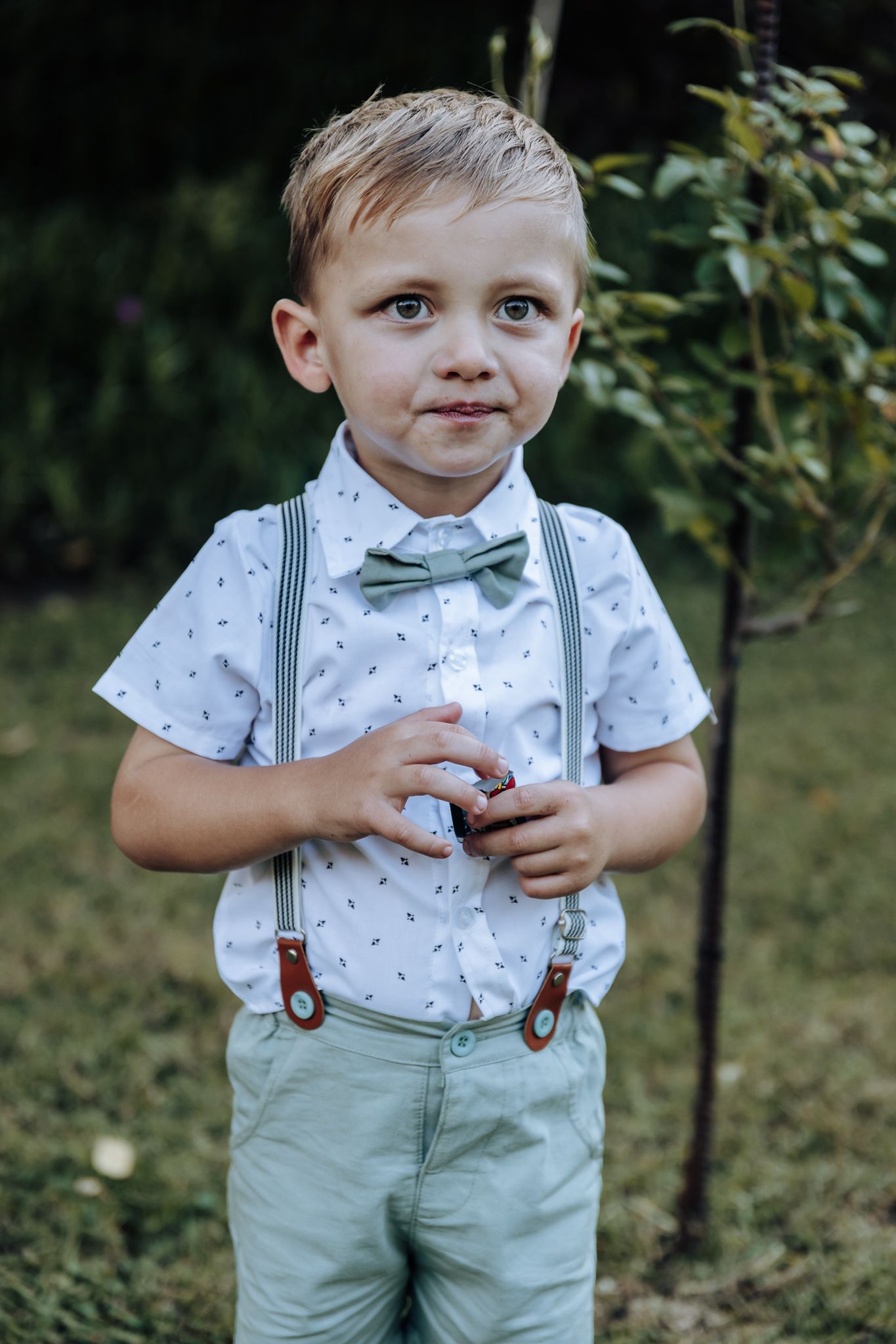 Young boy in dress clothes with suspenders and bow tie poses in garden.