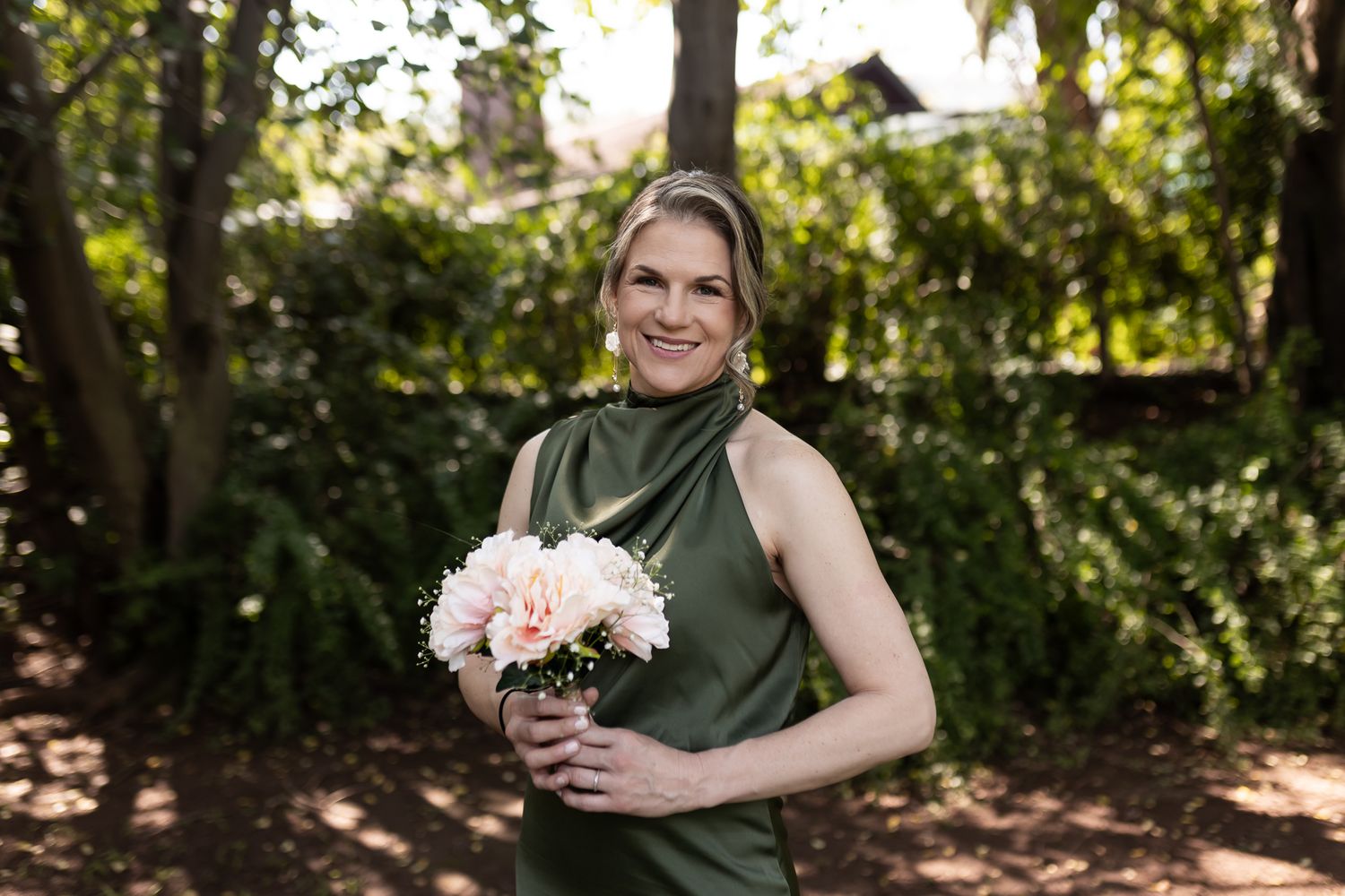 A person wearing a forest green halter dress holds pink flowers while standing in a garden.