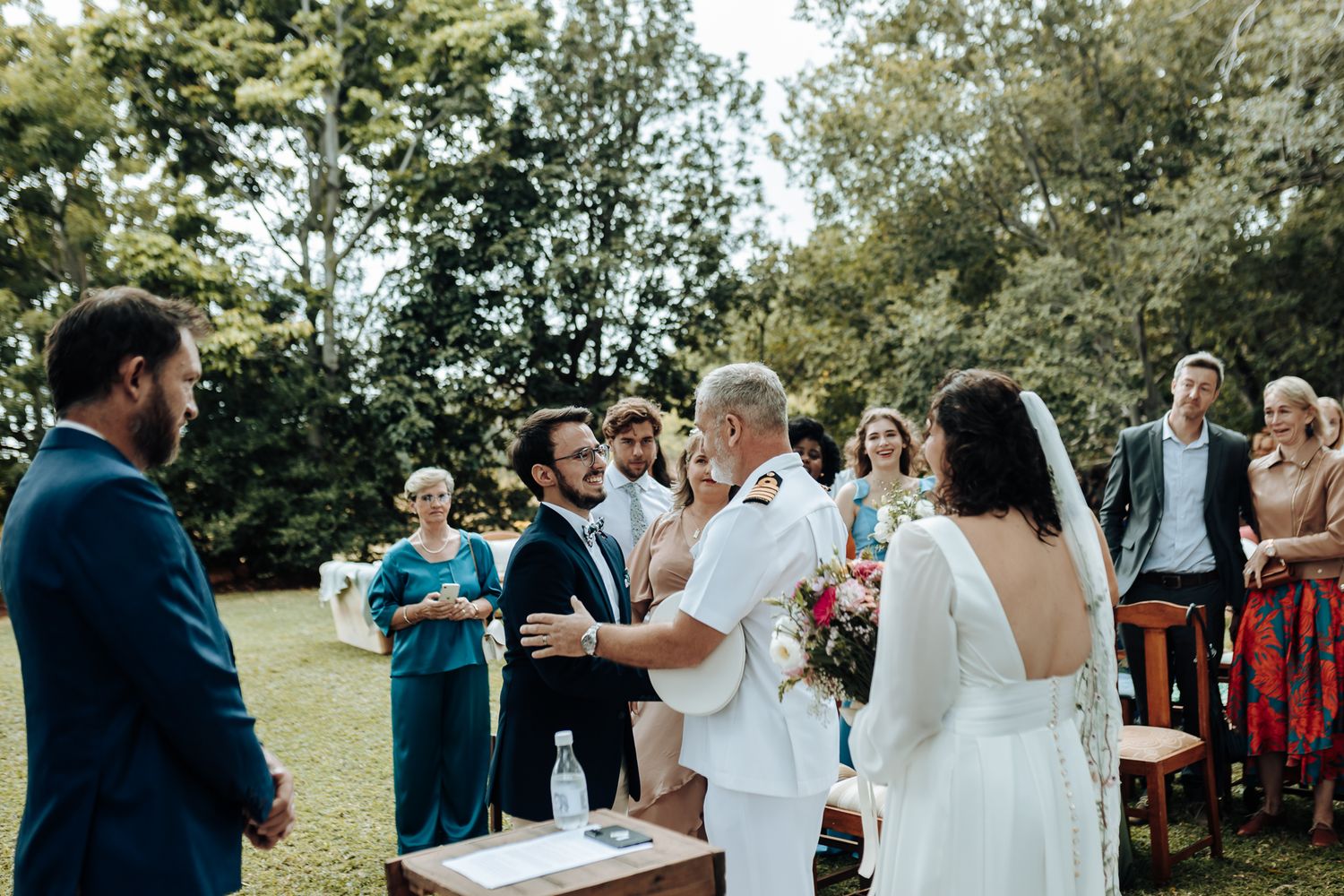 An outdoor wedding ceremony taking place beneath trees with guests gathered around a wooden podium.