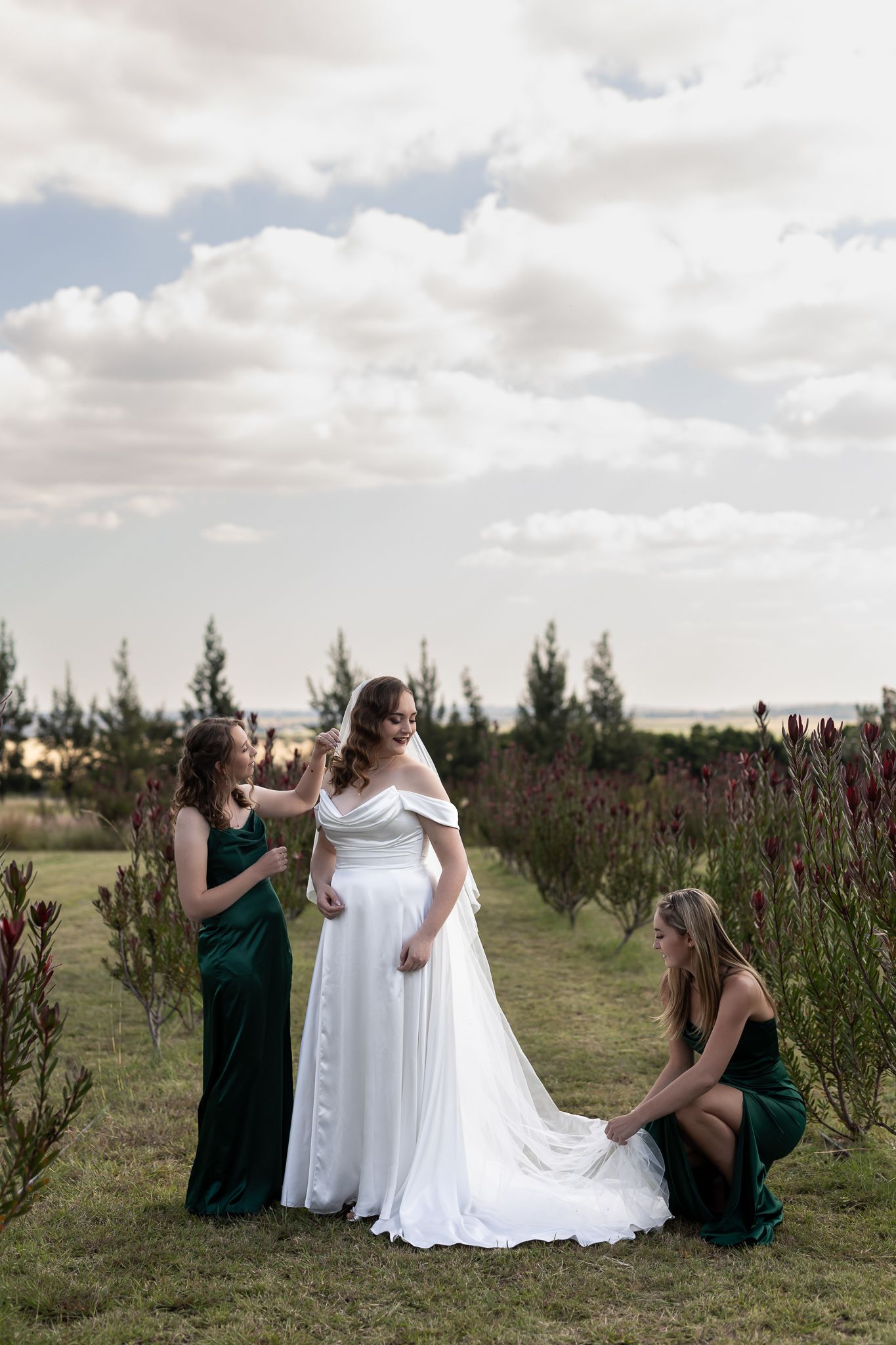 Wedding party gathered on grassy field helping adjust bride's dress among flowering plants.