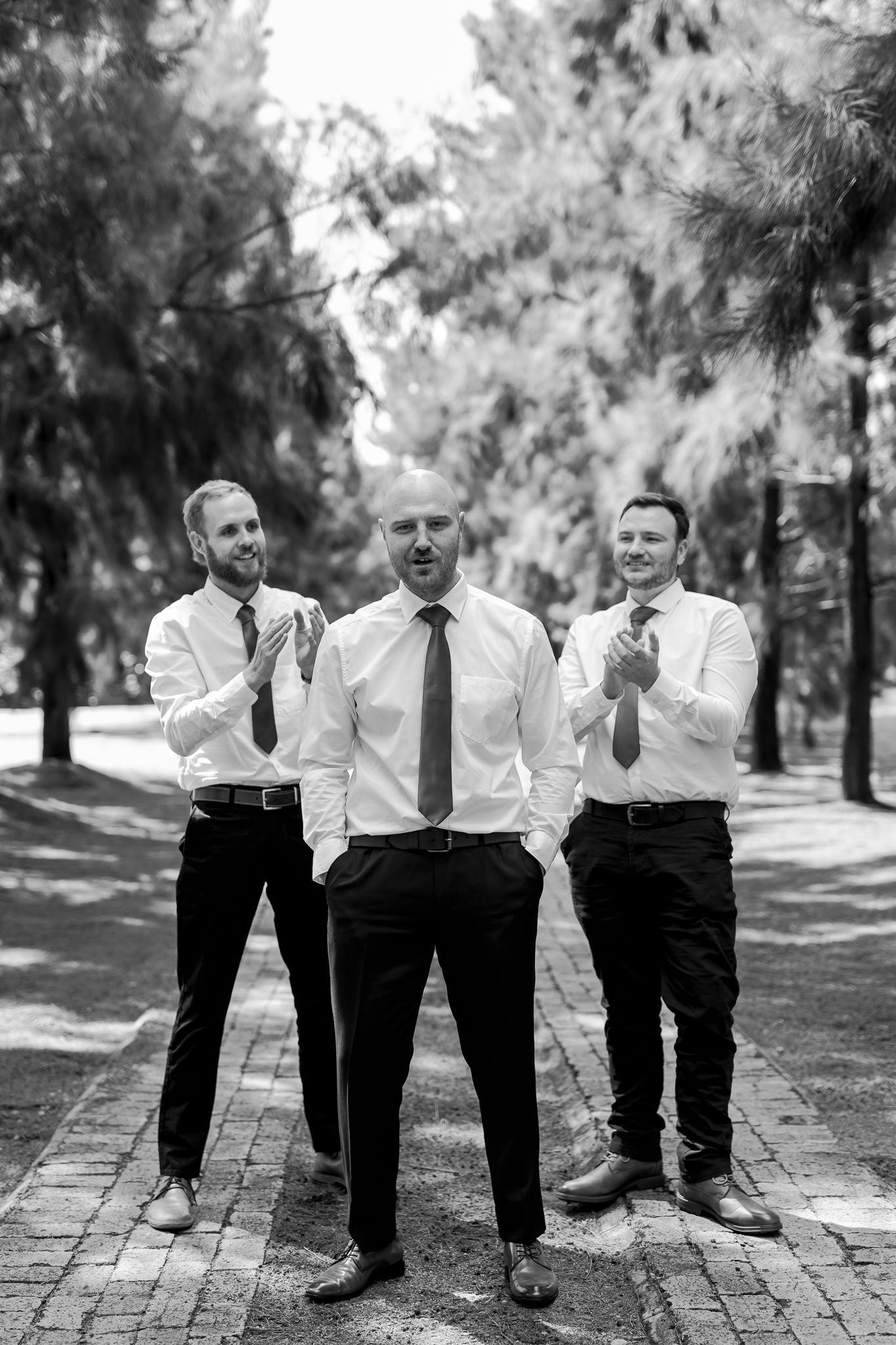 Three groomsmen in matching formal attire pose on a brick path in a black and white photo.