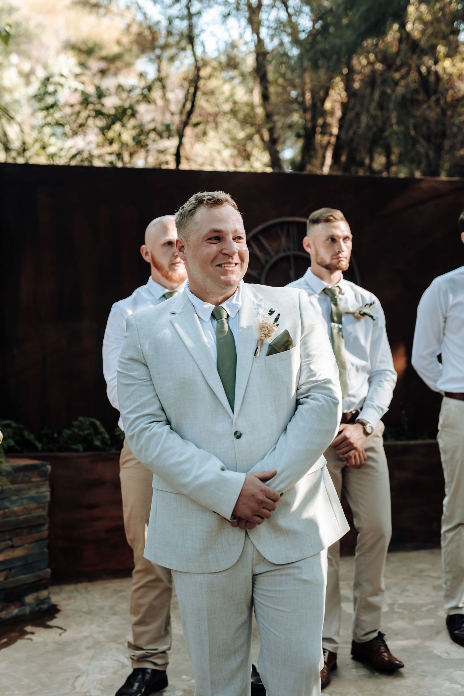 A groom in a light colored suit smiles during an outdoor wedding ceremony.