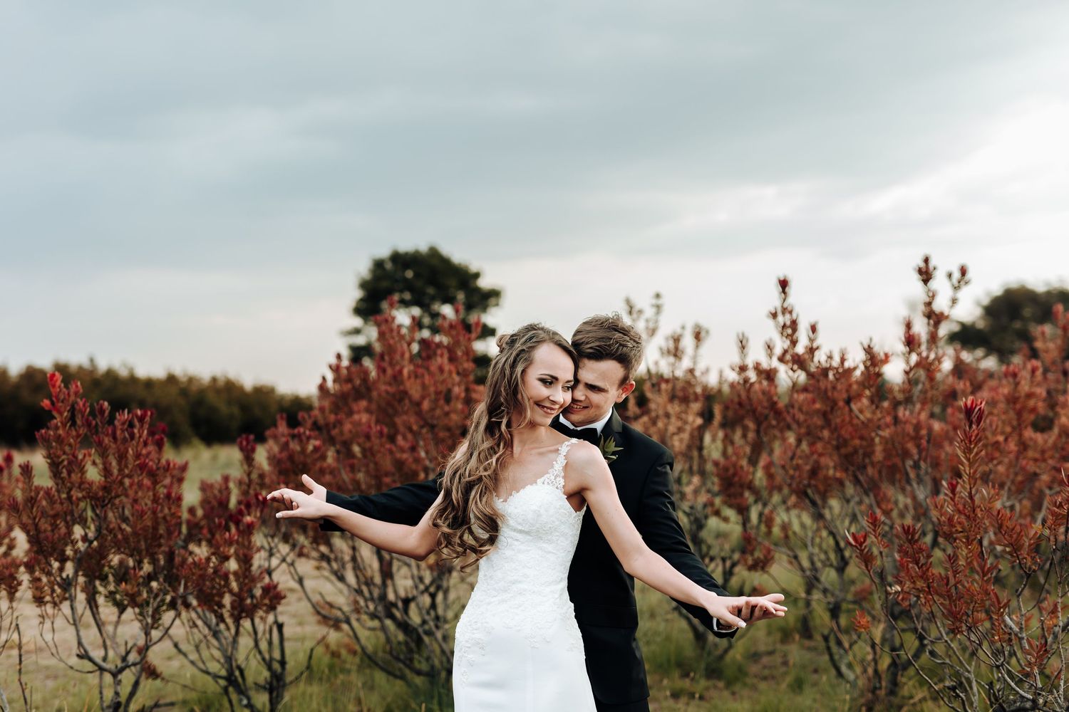 Joyful wedding couple poses playfully in field with autumn foliage.