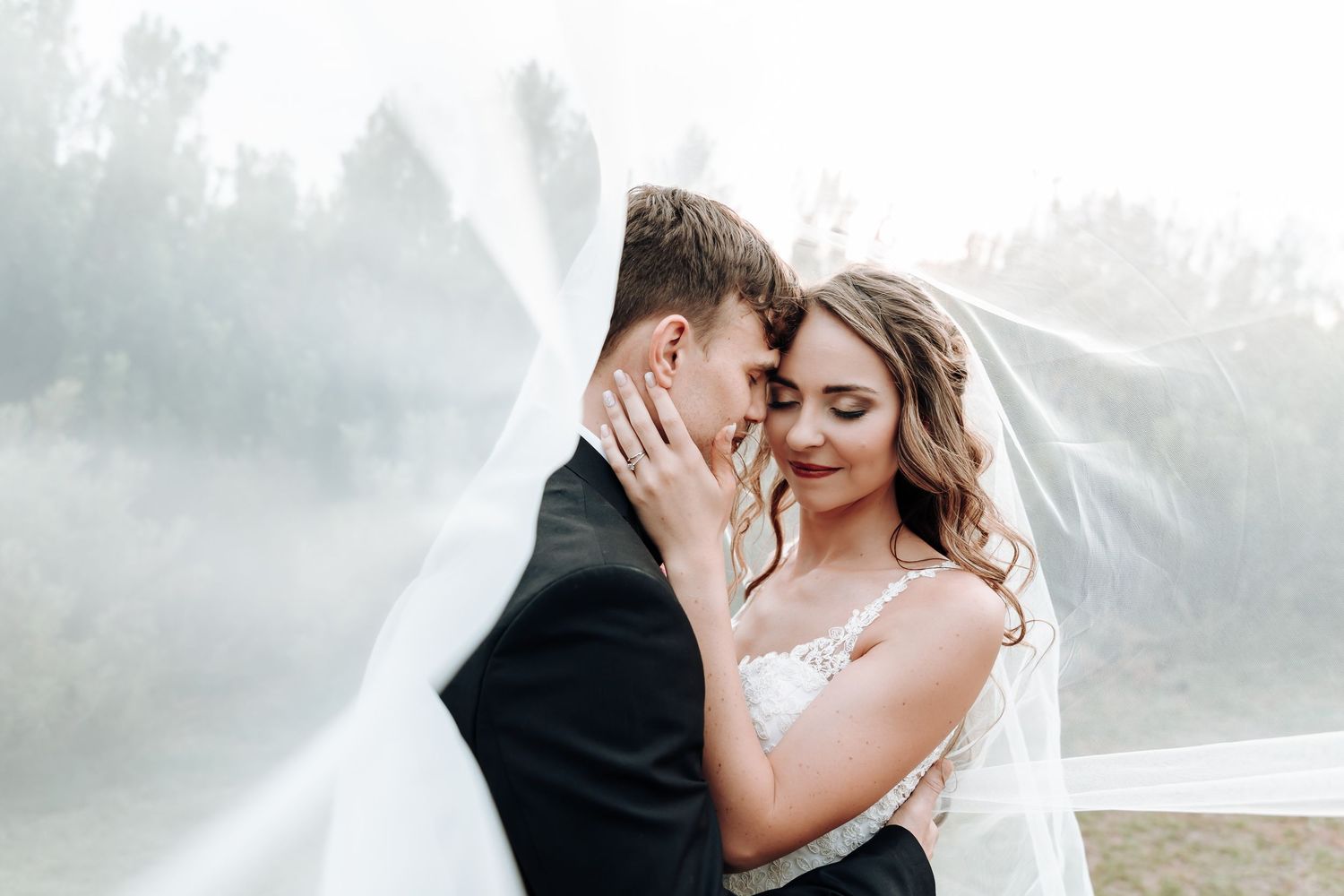 Romantic wedding couple embraces in ethereal foggy outdoor setting with flowing white veil.
