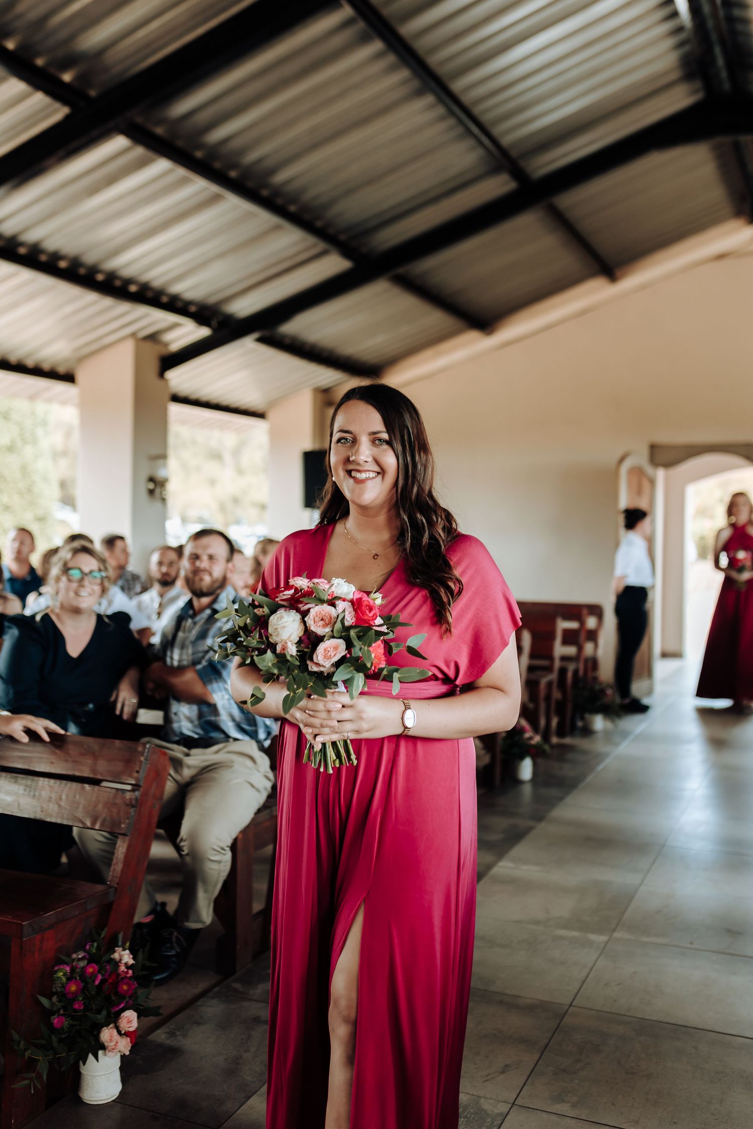 Bridesmaid walks down aisle in elegant red dress carrying pink floral bouquet.