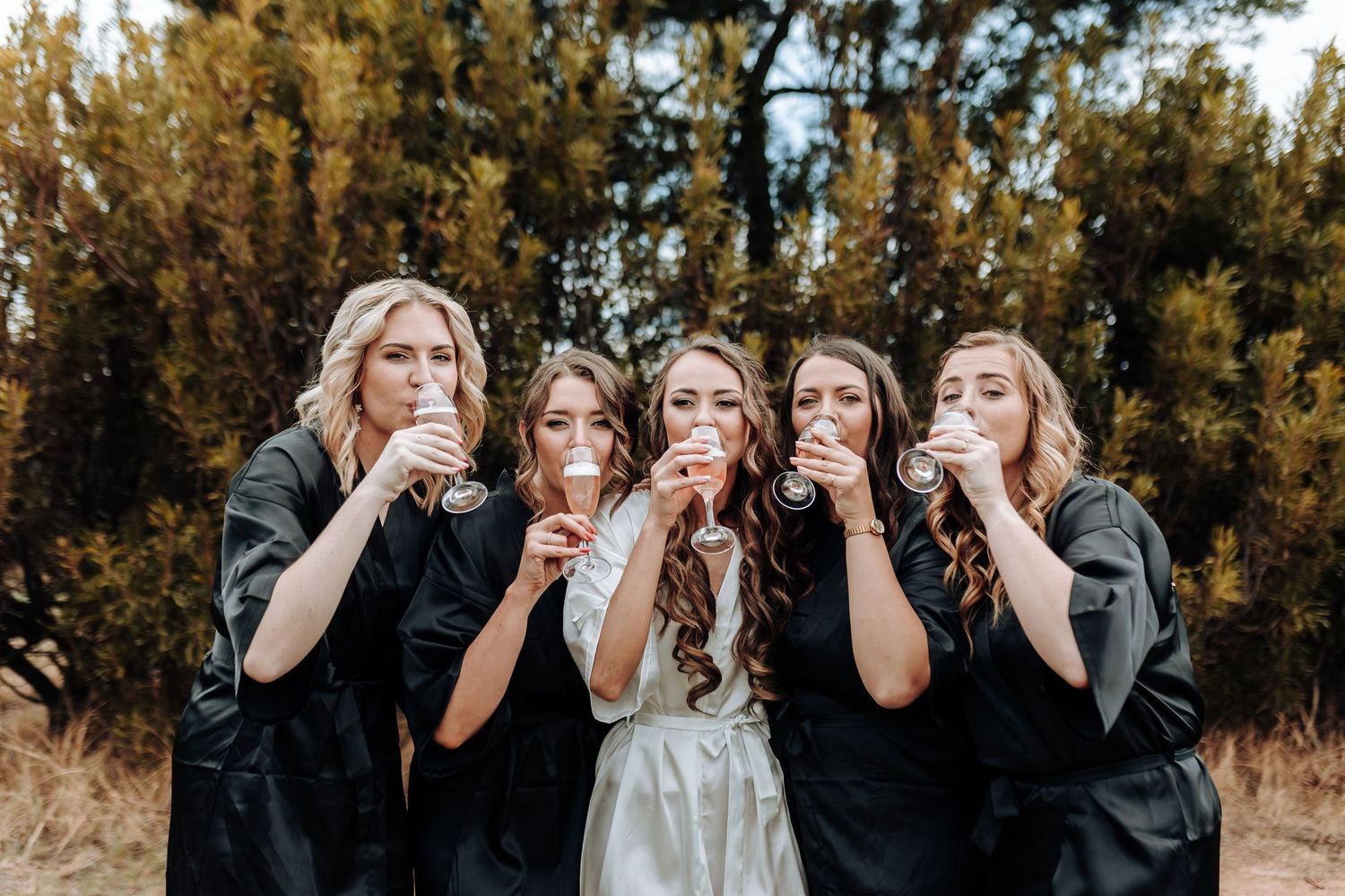 A bridal party in matching black robes shares a fun champagne toast moment against an outdoor pine tree backdrop.
