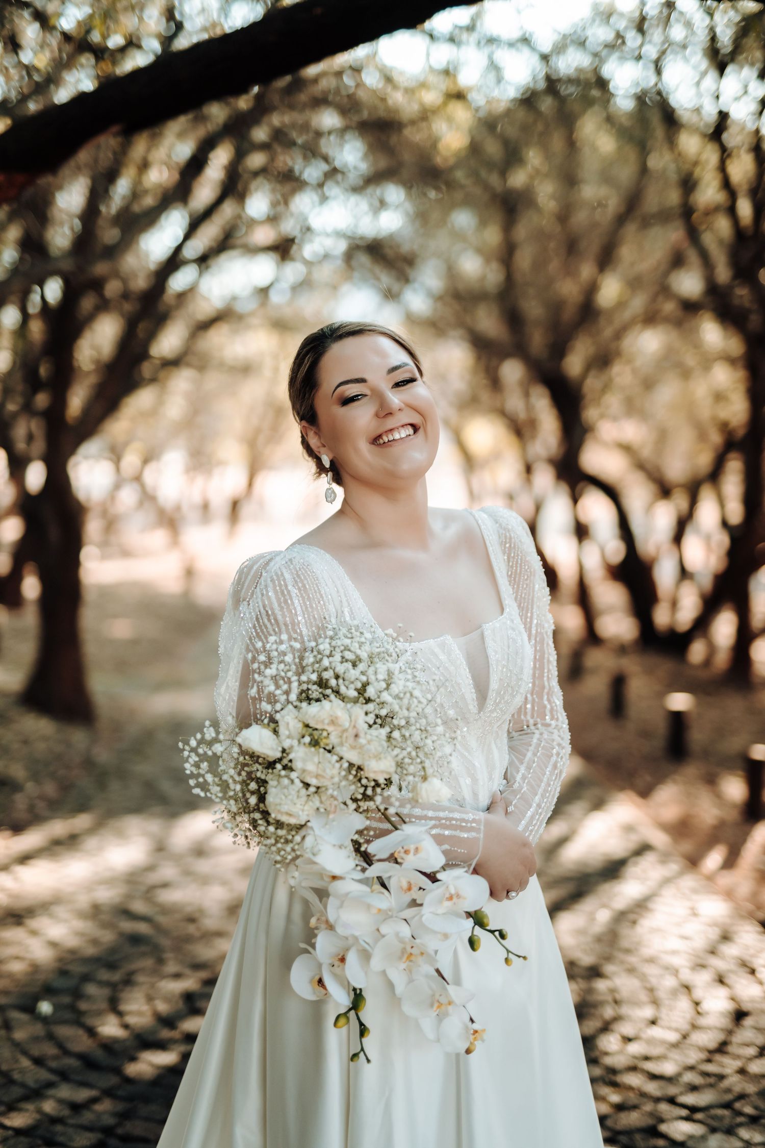 Radiant bride smiles while holding white floral bouquet in sunlit forest setting.