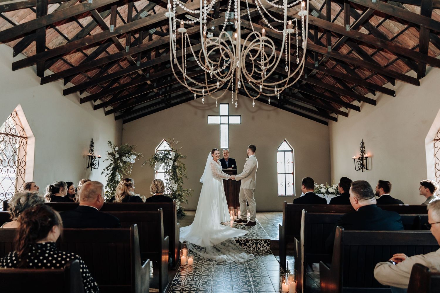 Wedding ceremony under wooden beams and chandelier in intimate chapel with arched windows.