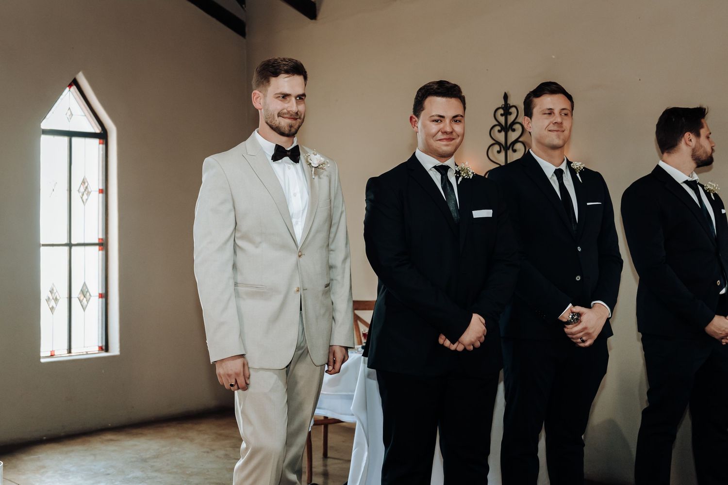 Groom with groomsmen in mix of light and dark suits stand in chapel.