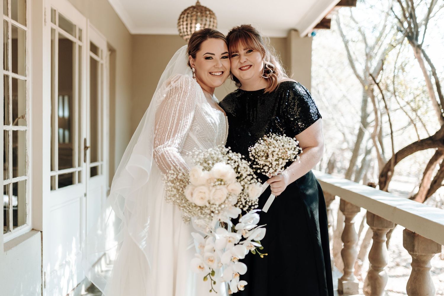Bride in white wedding dress poses with woman in black on elegant porch with white floral bouquet.