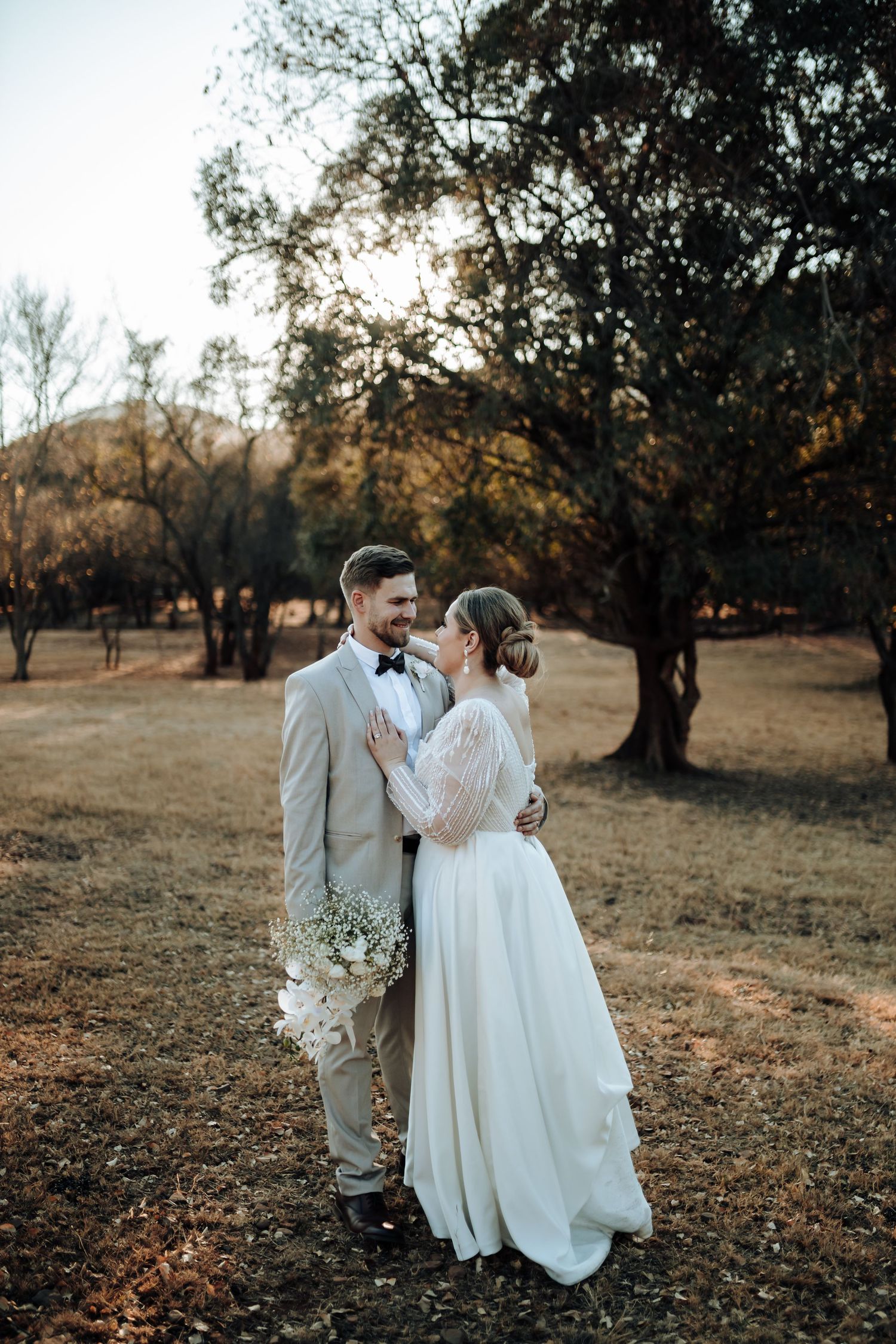 Sunset wedding photo with couple standing among trees holding delicate white bouquet.