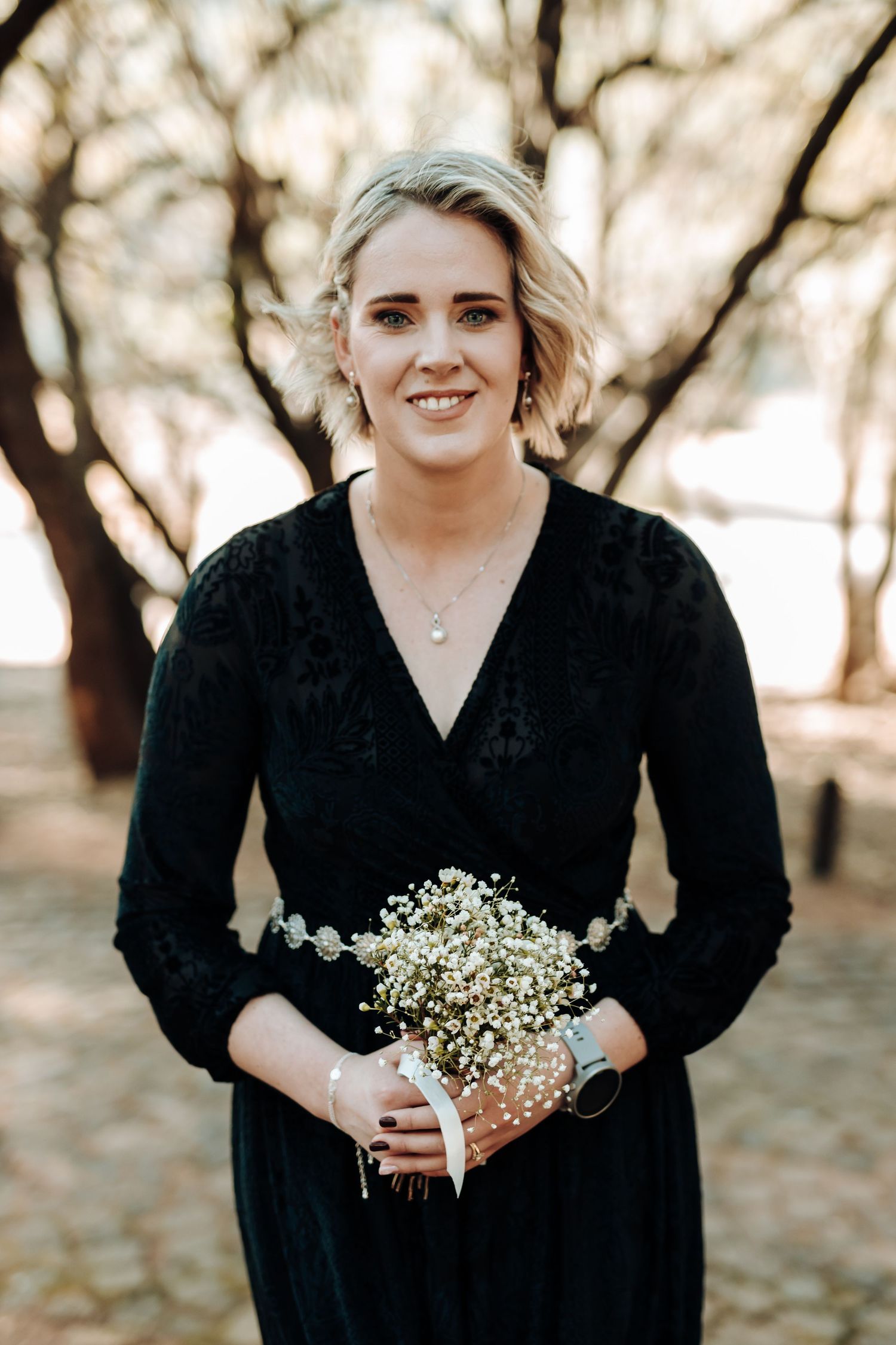 Bridesmaid in black v-neck dress poses with white bouquet against autumn trees.