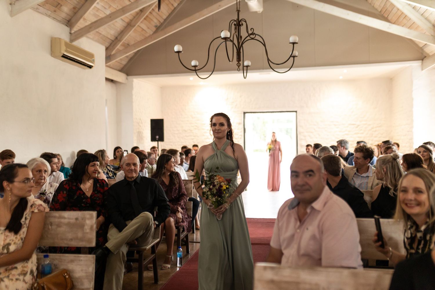 Wedding party member proceeds down aisle in sage dress while guests look on in chapel setting.