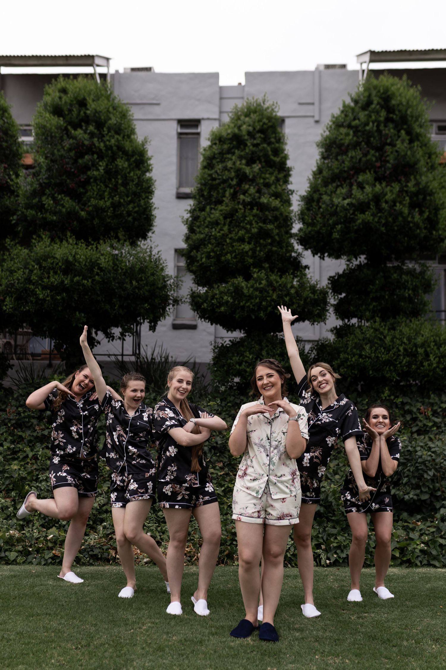 A group in matching floral pattern outfits strikes fun poses outdoors in front of trees and a building.