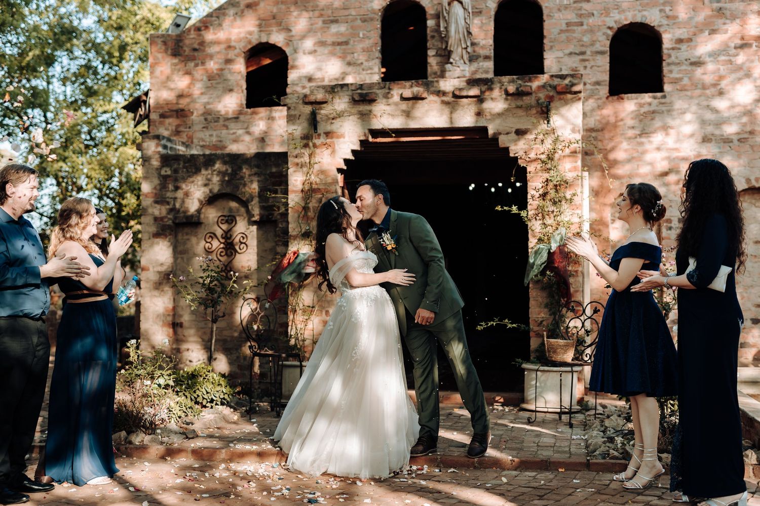Wedding couple kisses outside historic brick building while guests celebrate with confetti.