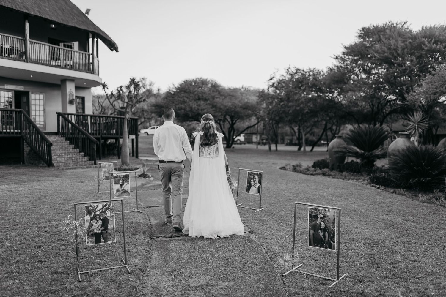 Wedding couple walking past framed photos displayed on stands in a garden.