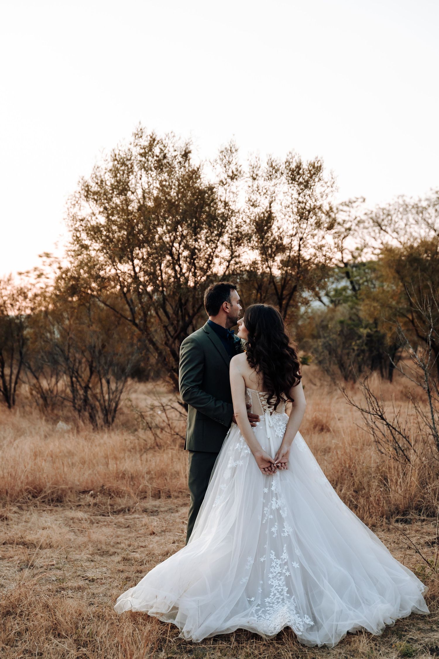 Two newlyweds sharing an intimate moment in a rustic outdoor setting with dried grass and trees at sunset.