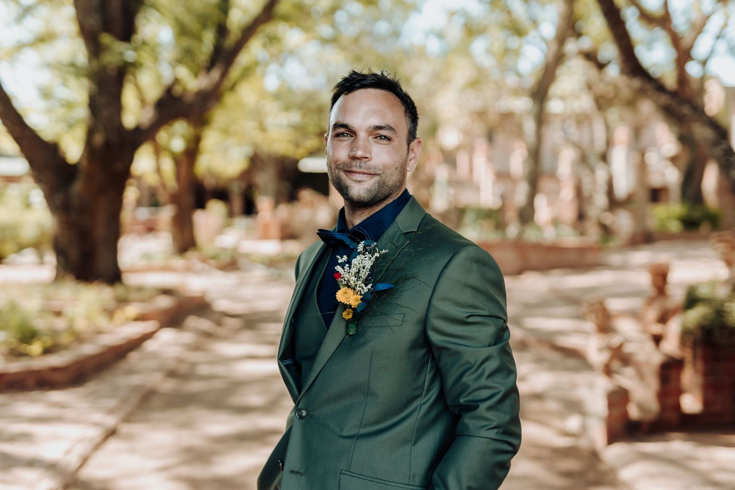 A person in a formal dark green suit with boutonniere poses in a sunny outdoor garden setting.