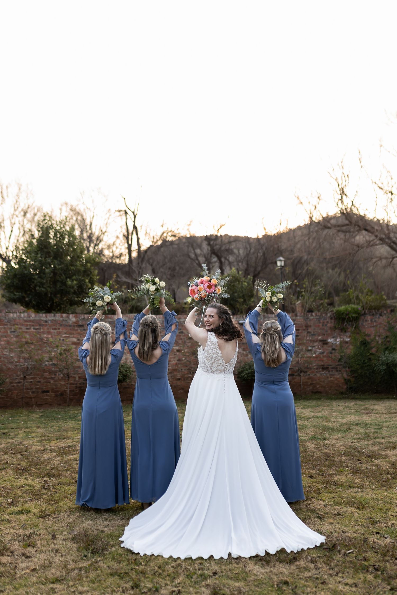 A bridal party in blue bridesmaid dresses and white wedding gown poses with their backs turned on a rustic outdoor lawn.