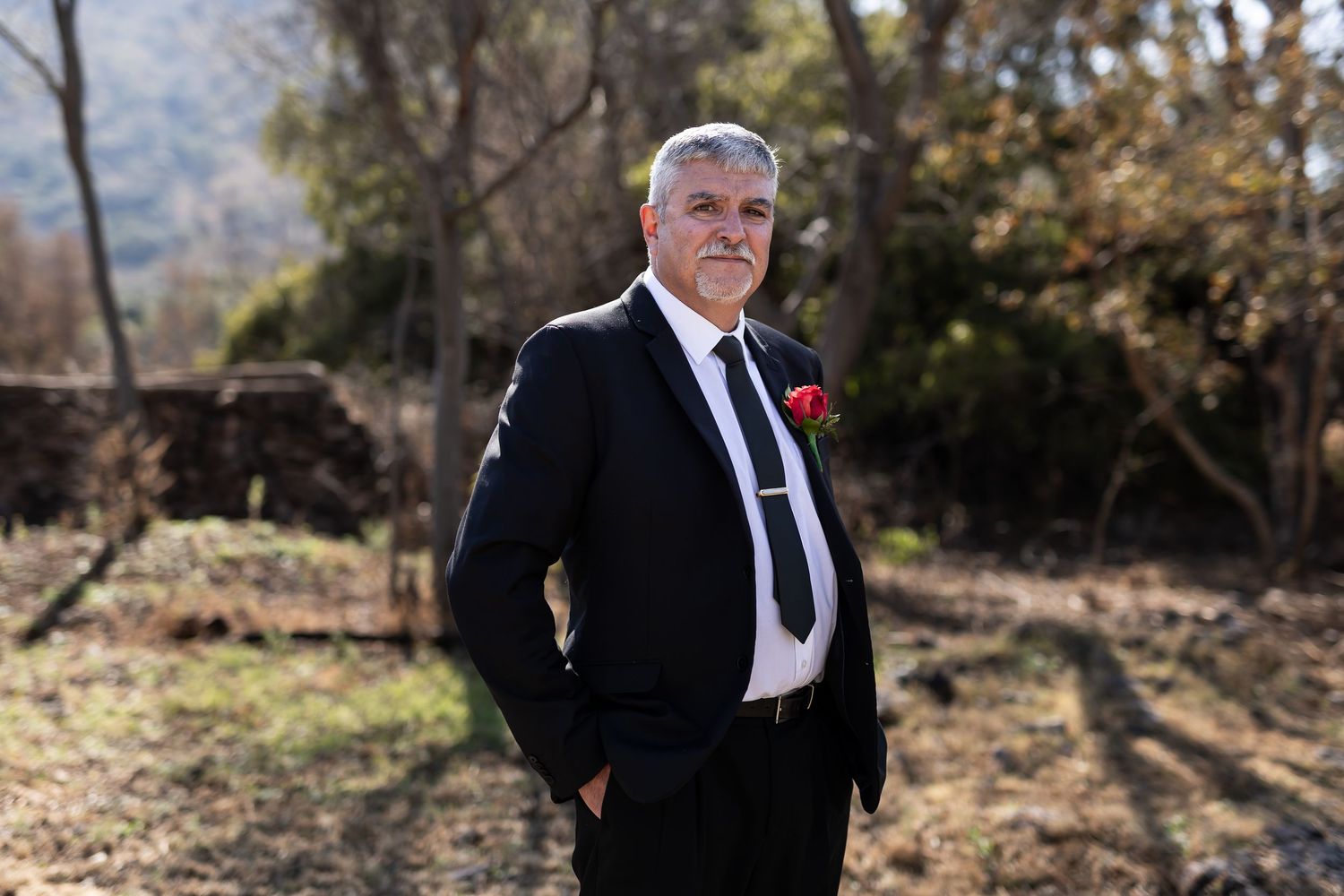 A distinguished person in a black suit with red boutonniere poses outdoors in natural lighting with trees in background.