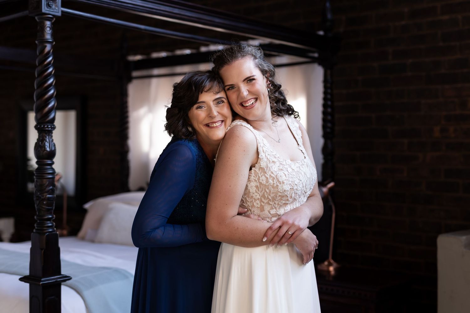 Two smiling figures embrace while standing in a bedroom, one wearing a navy dress and one in a white lace wedding gown.