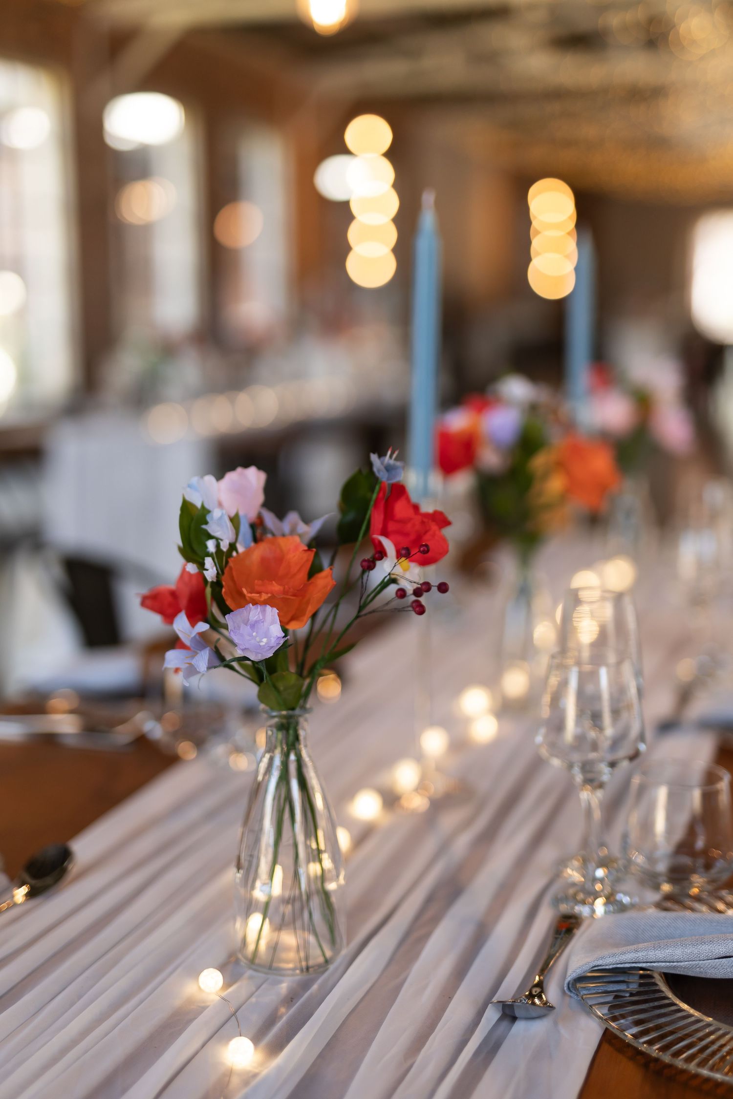 Elegant table setting with small floral centerpiece featuring red and pink blooms, blue candles and bokeh lighting in background.