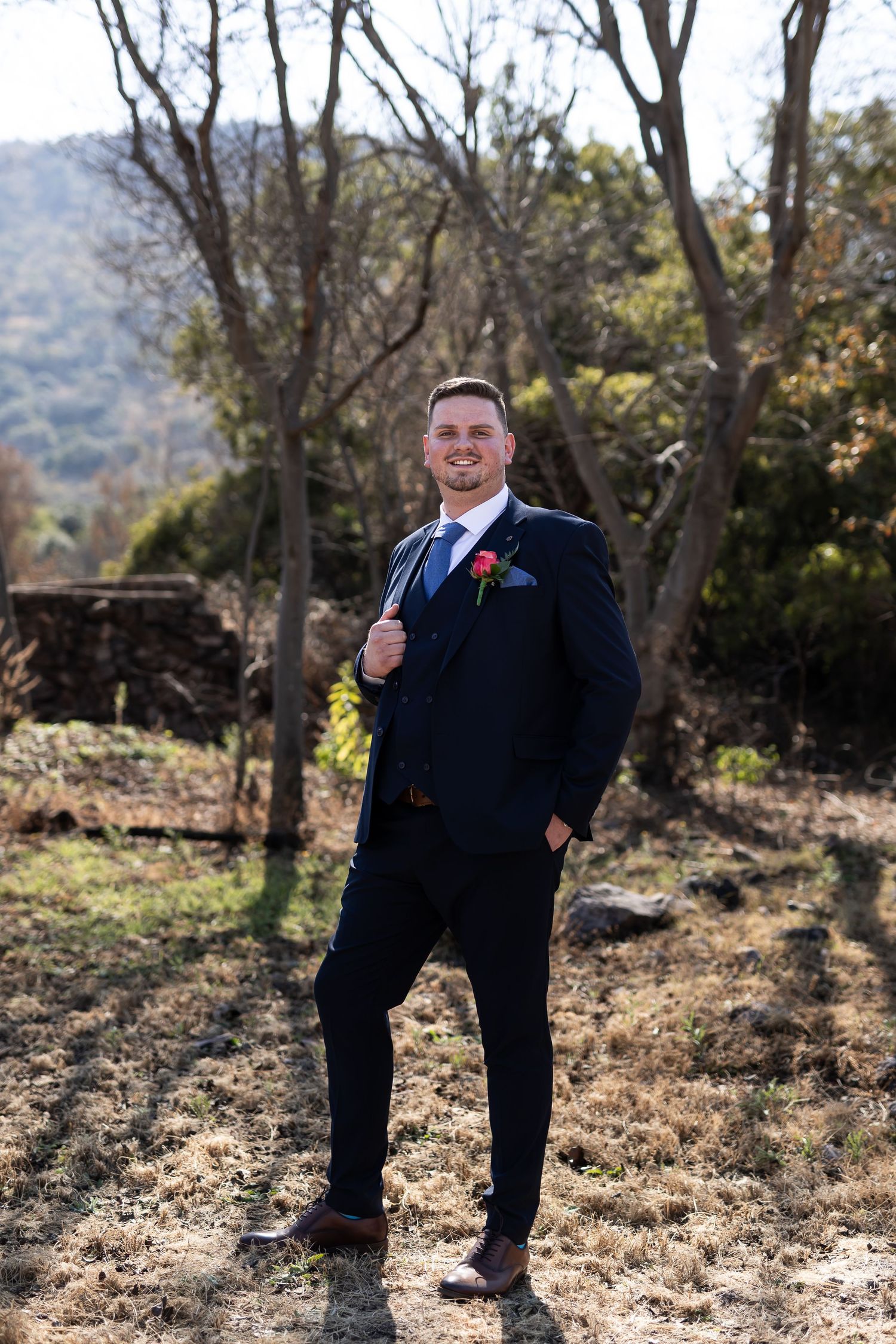 A person in a navy blue suit and tie stands in an autumn landscape with trees and hills in the background.