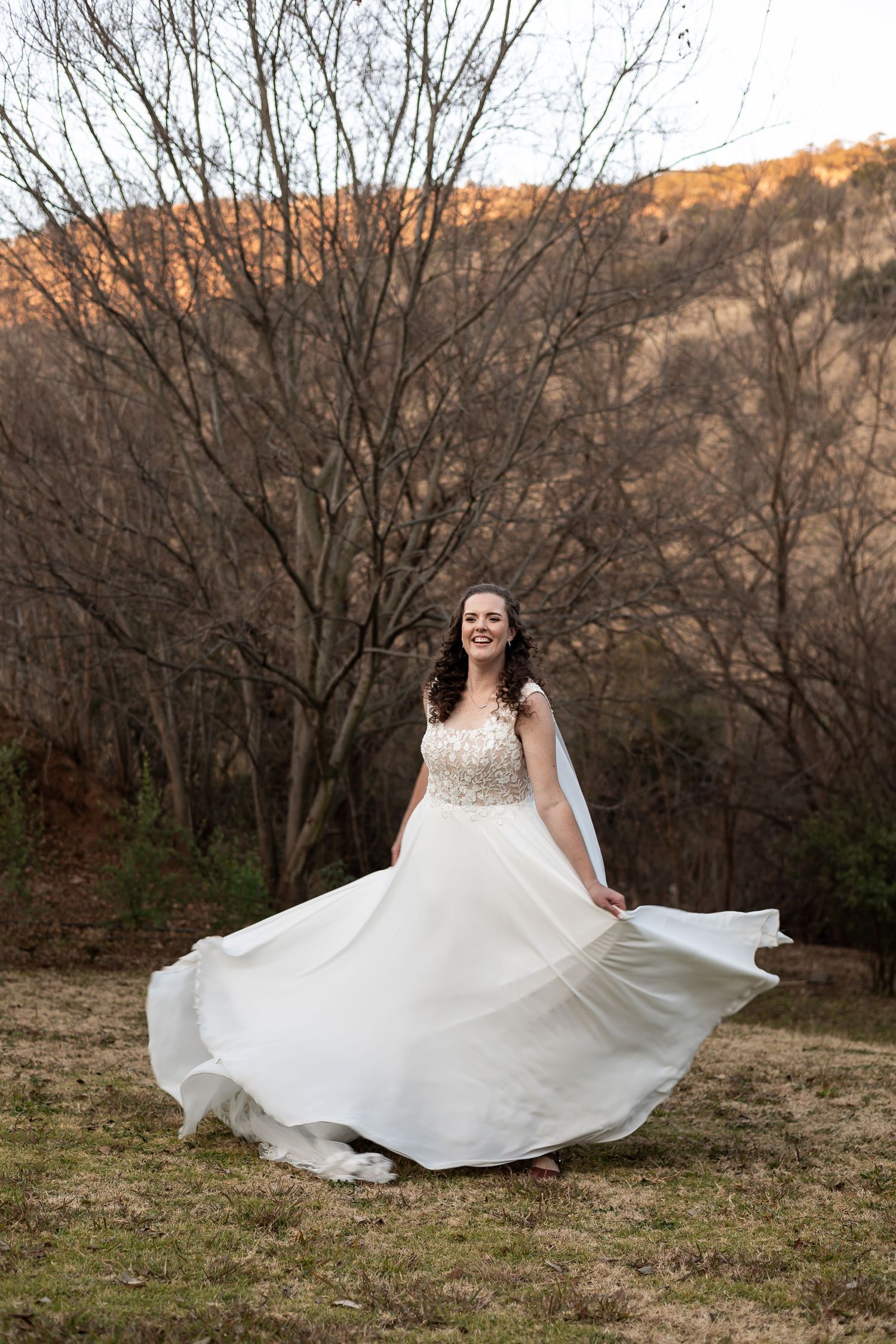A bride in a flowing white ballgown twirls joyfully in a rustic outdoor setting at sunset.