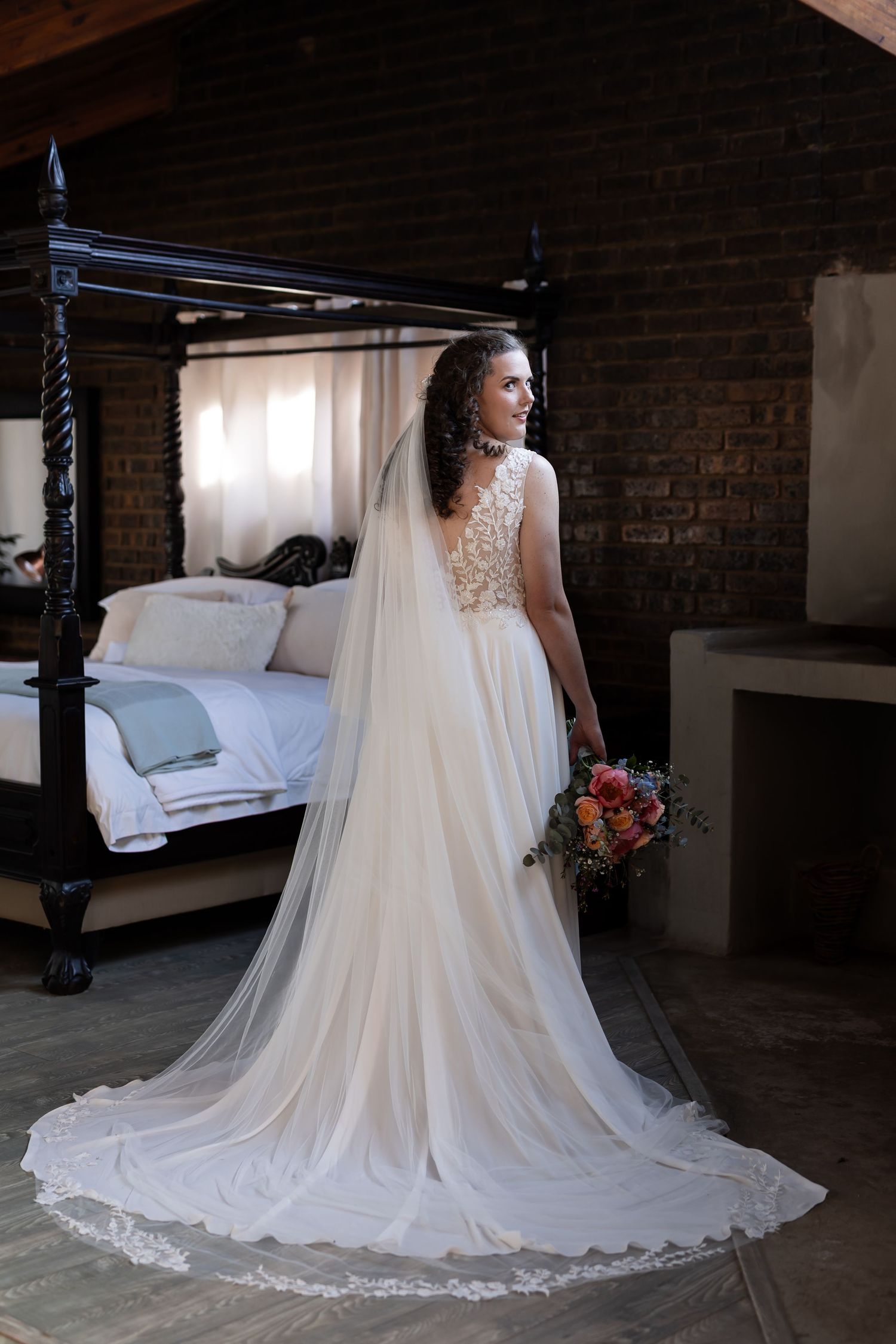 A bride in a flowing white lace wedding gown with a dramatic cathedral train stands in a rustic brick room by a window.